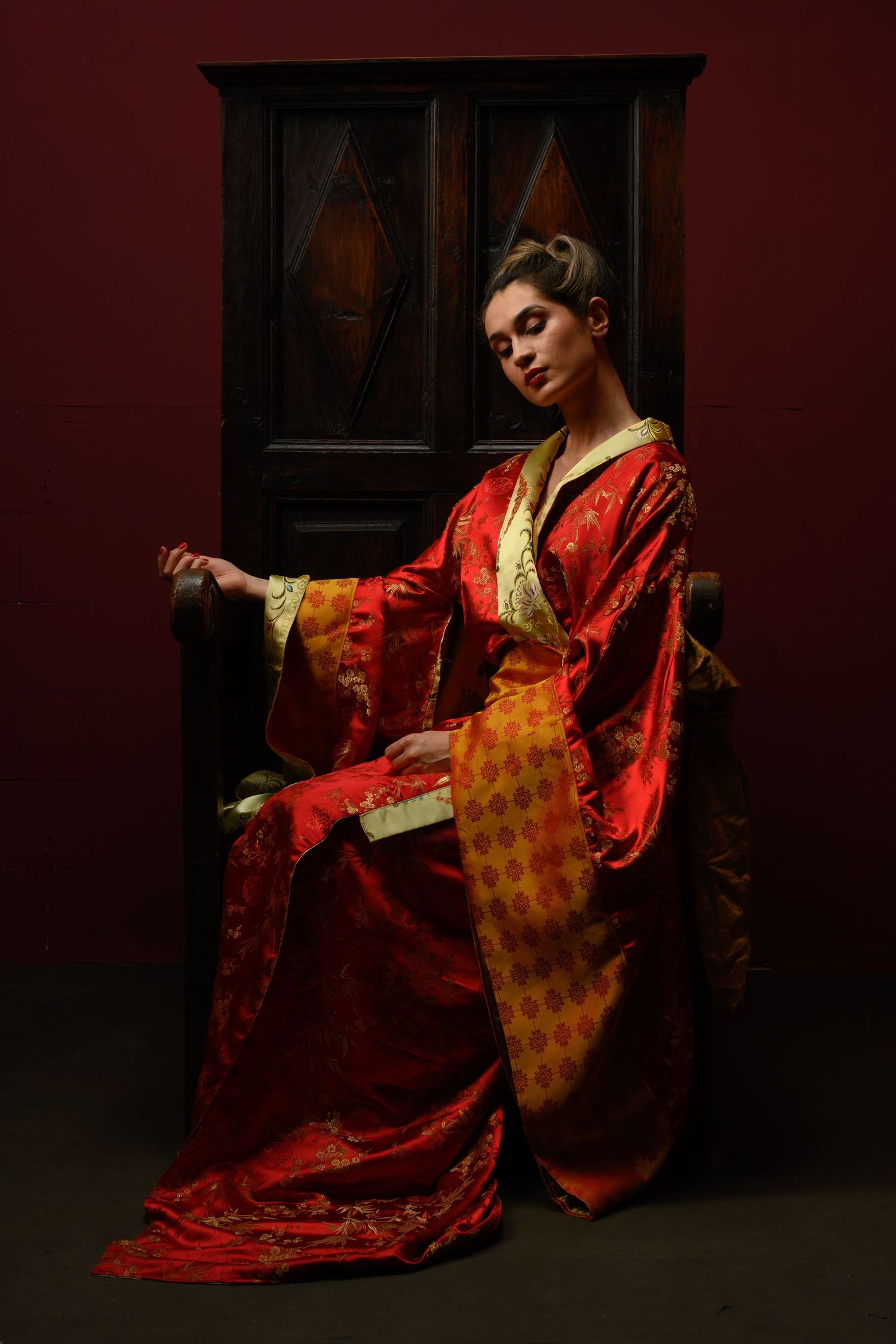 Dramatic portrait of a person in luxurious red and gold traditional Asian robes against dark background.