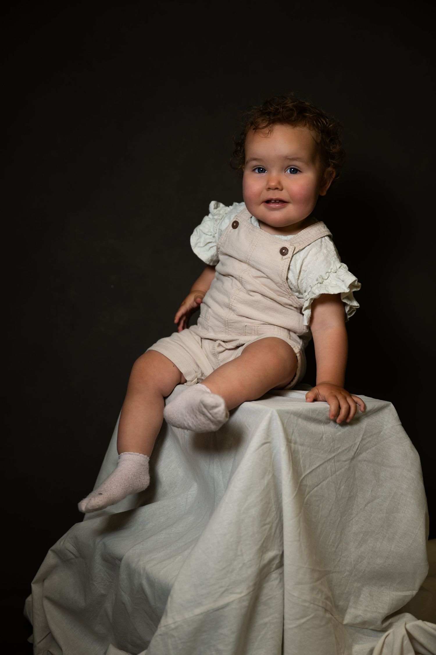 Toddler in vintage-style outfit sitting on draped white fabric.