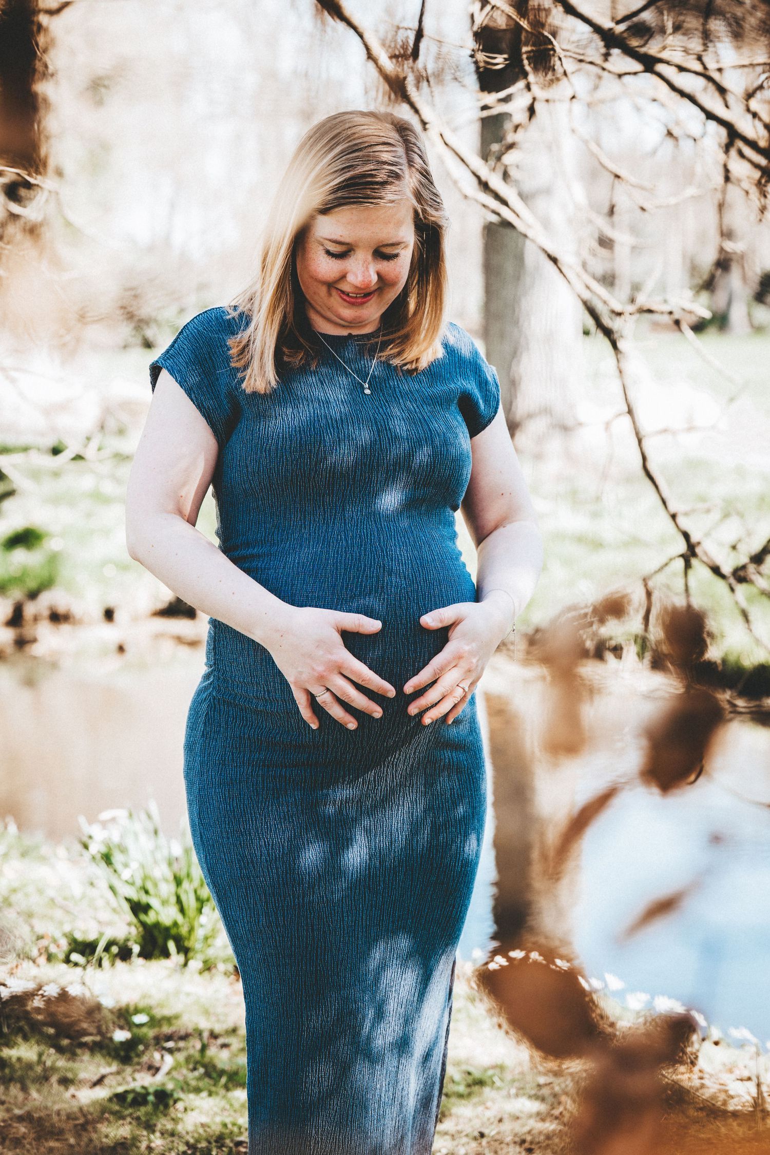 Maternity photoshoot in a blue dress under blooming spring trees with dappled sunlight.