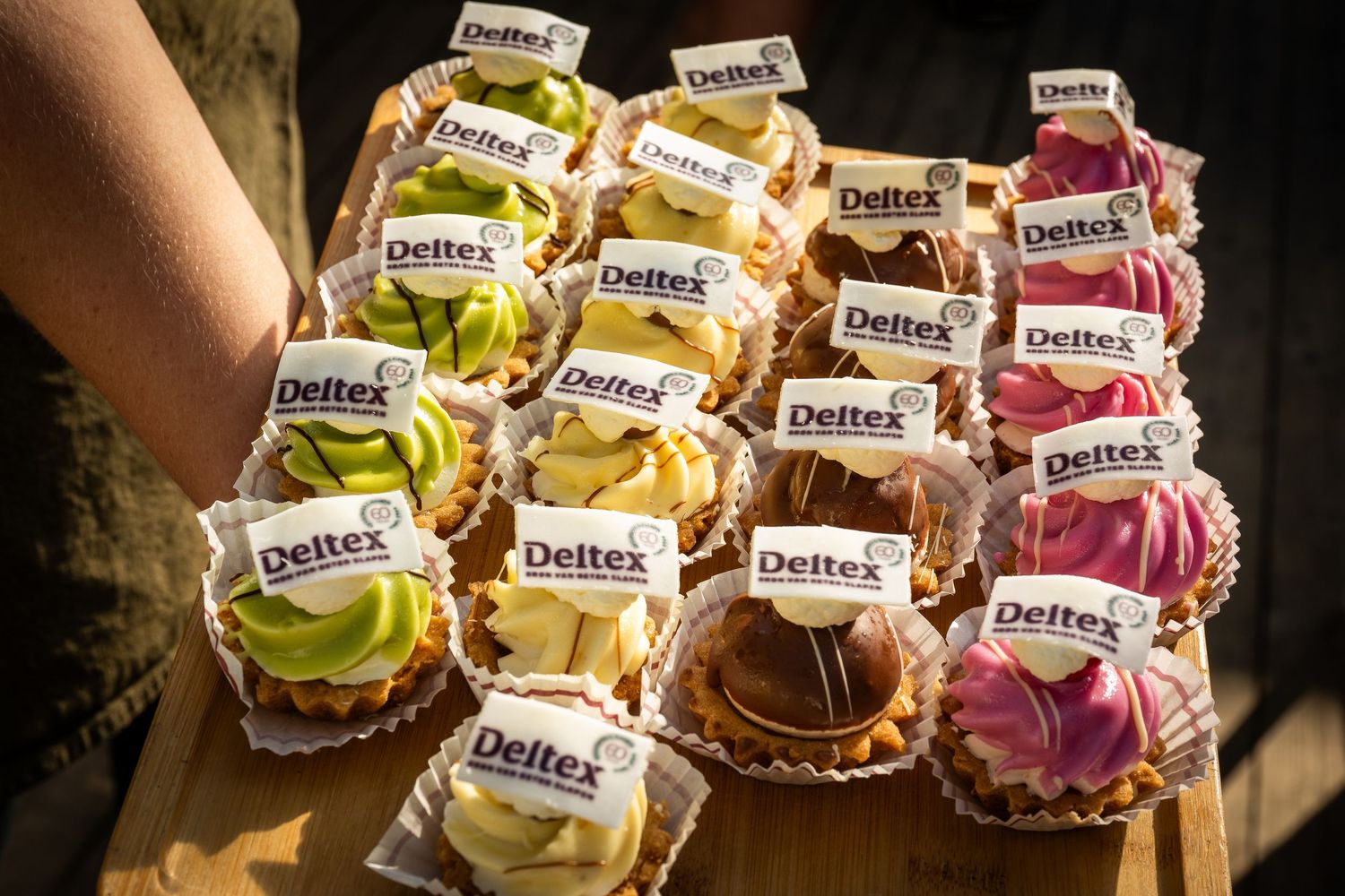 Colorful cupcakes topped with pink and yellow frosting and decorated with Zweifel brand flags in a display case.