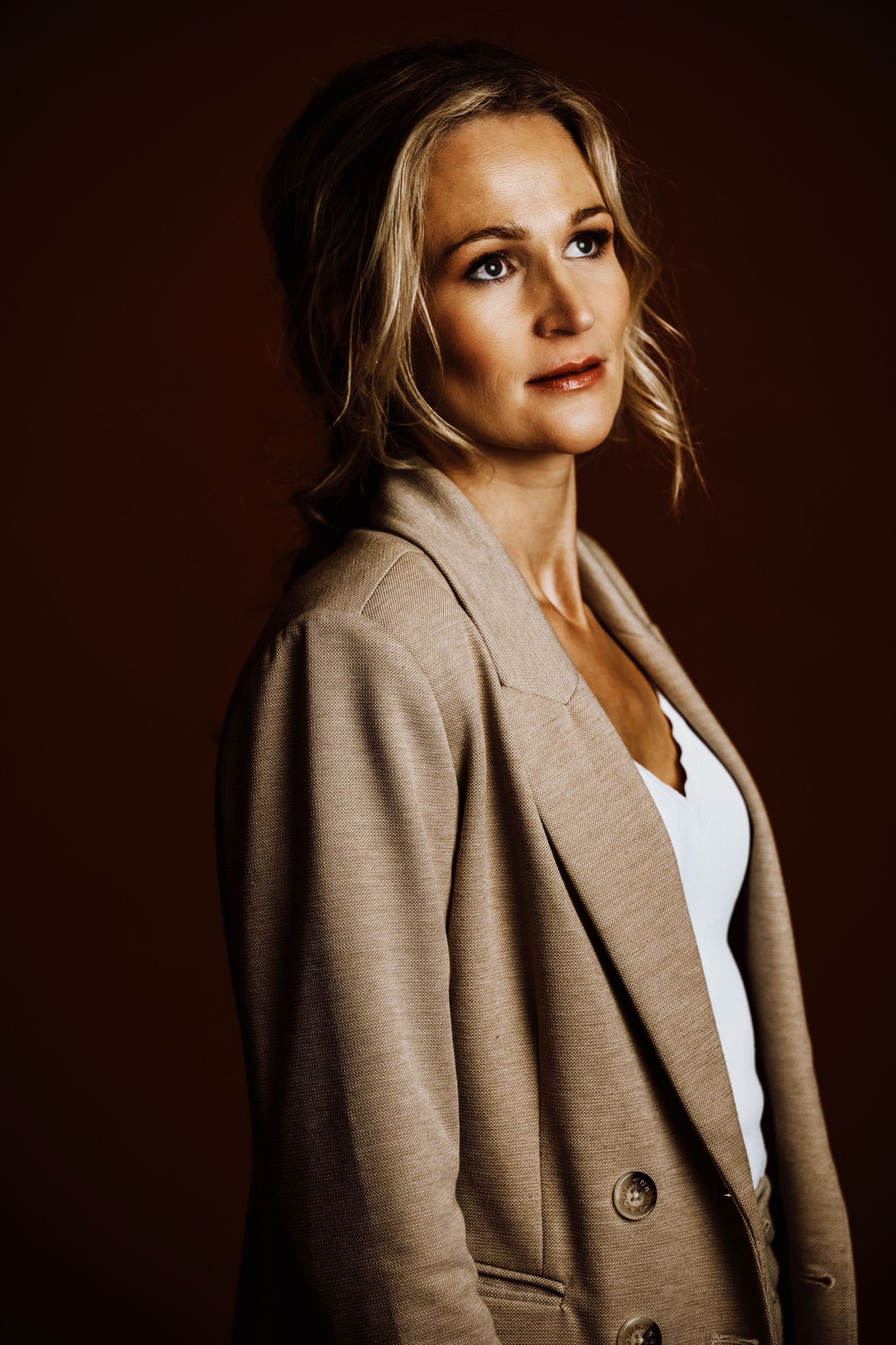 Professional portrait of a person in a beige blazer and white top against a dark brown background.
