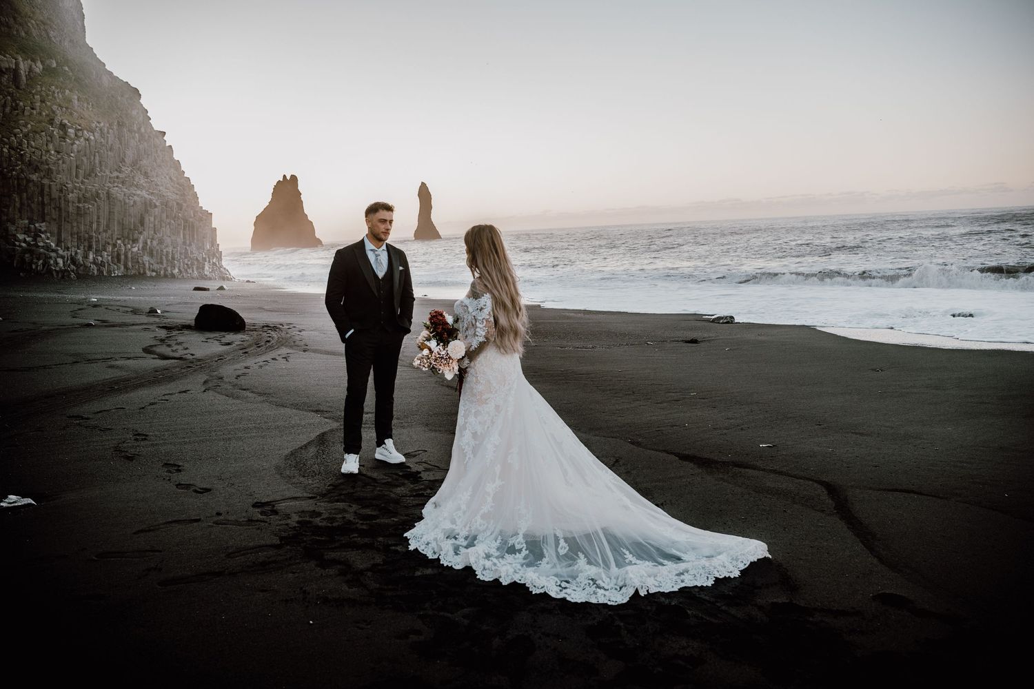A couple walks along a dramatic black sand beach at sunset with ocean waves and cliffs in the background.