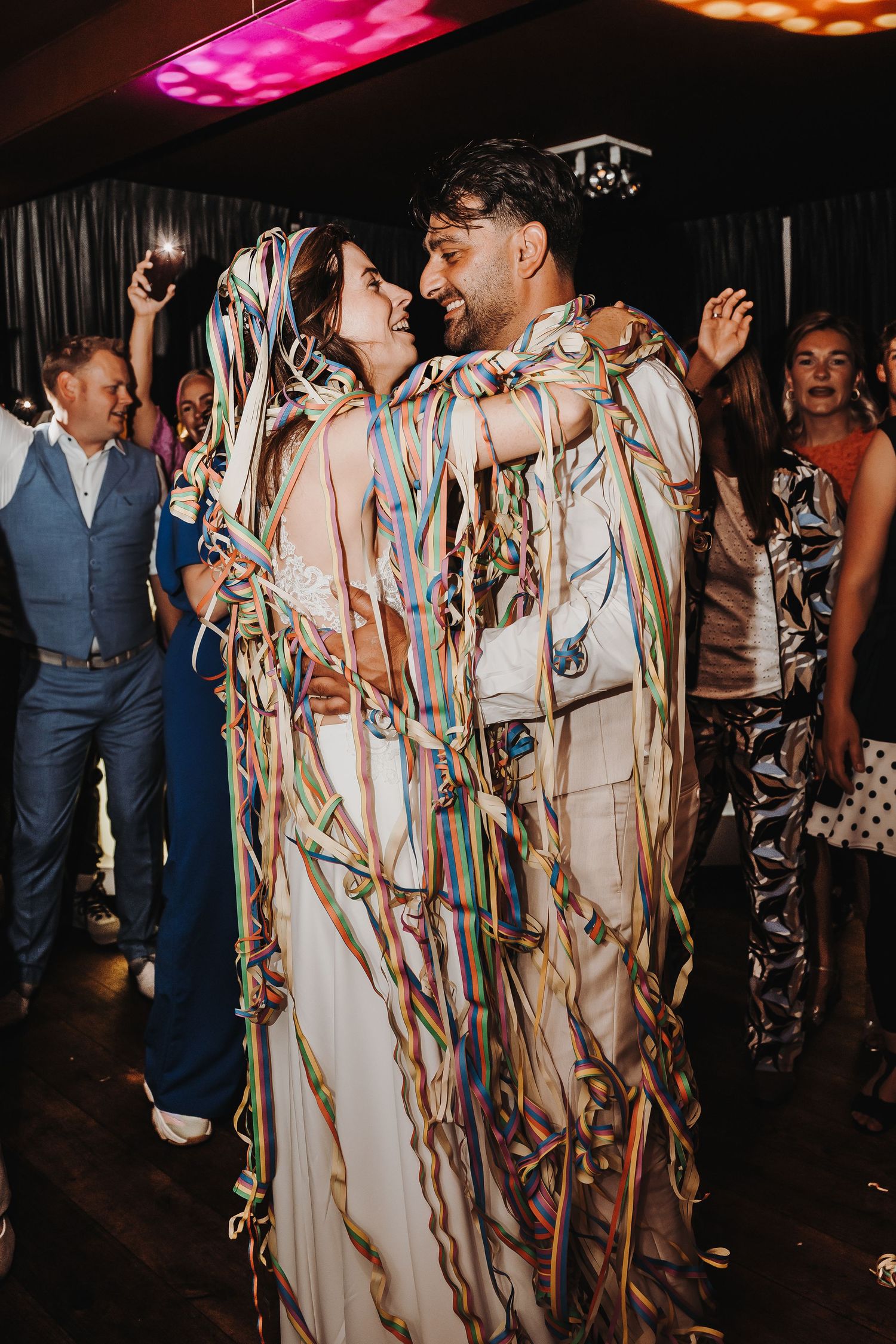 Wedding party dancing and celebrating with colorful streamers in a dark reception venue.