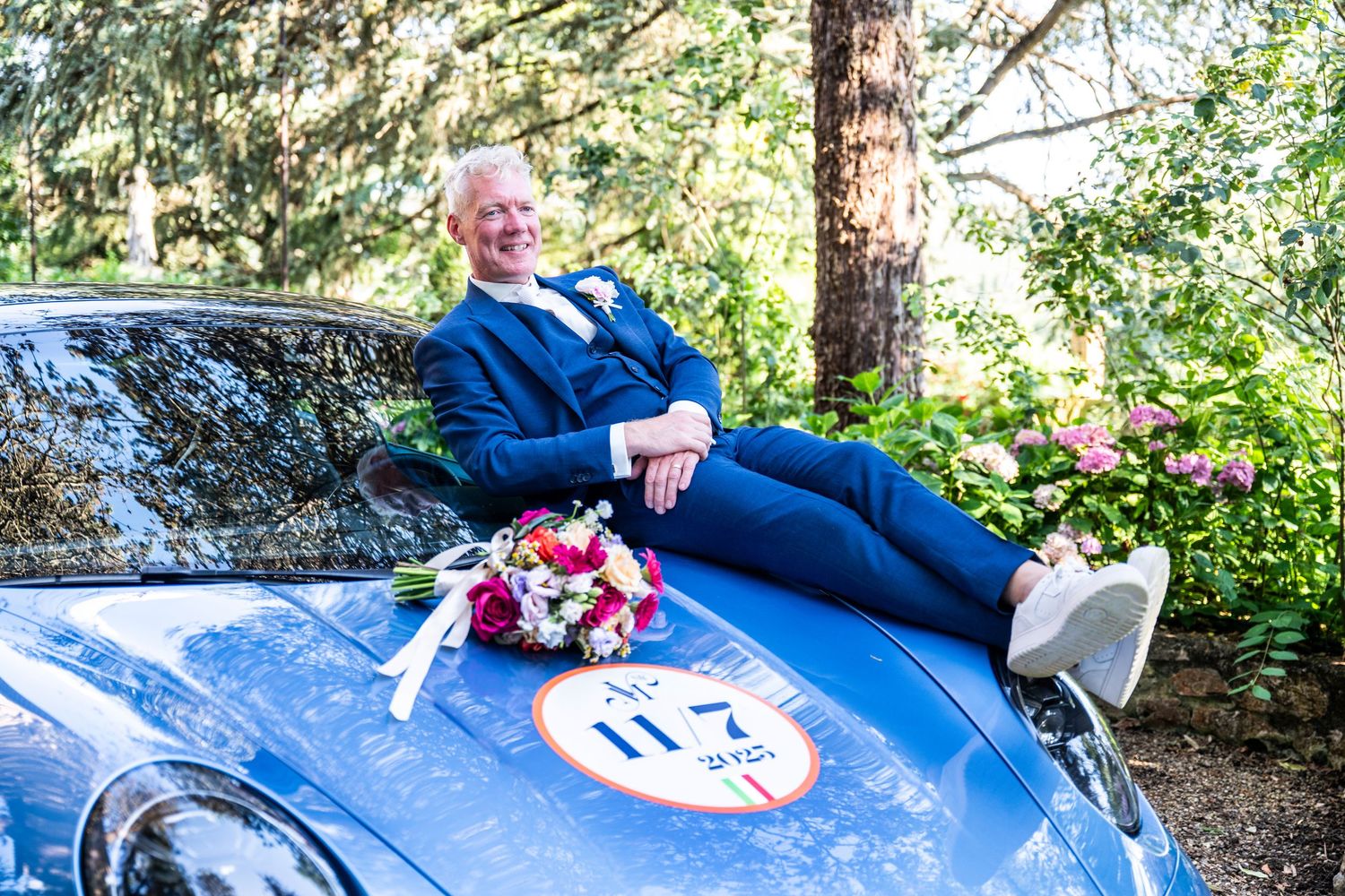A person in blue suit reclines on vintage blue car with wedding bouquet.