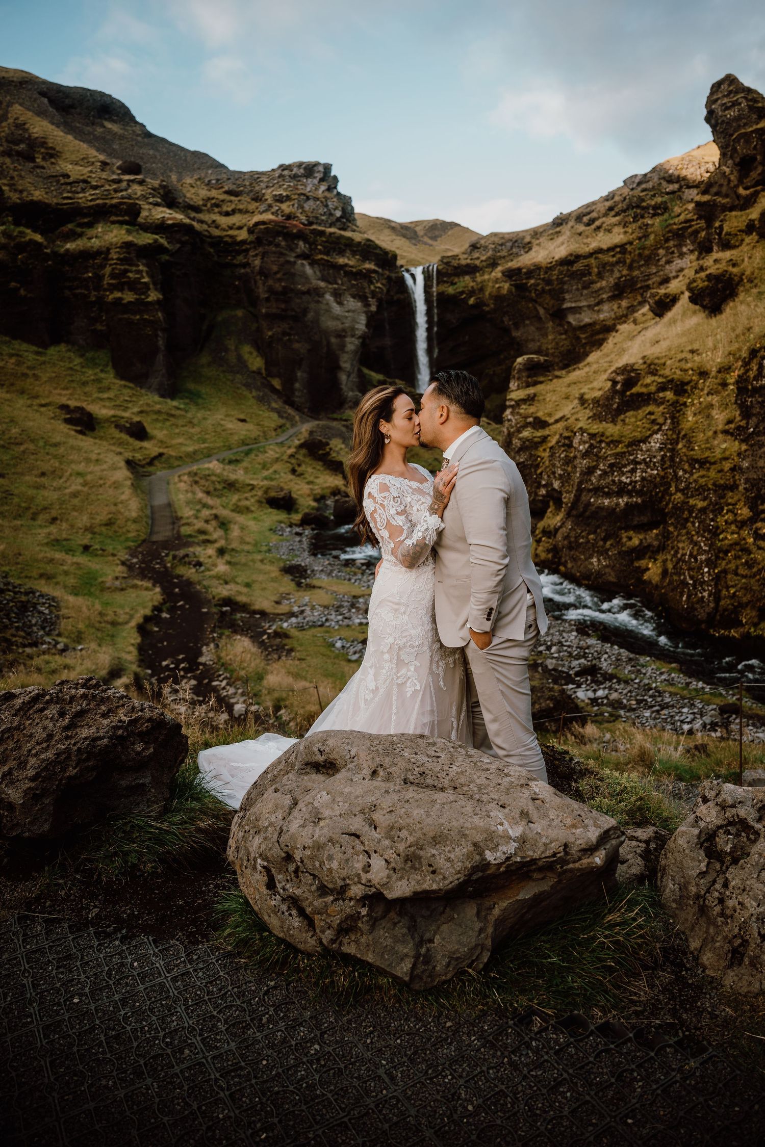 A couple shares an intimate moment on a large boulder overlooking a majestic mountain landscape with waterfalls.