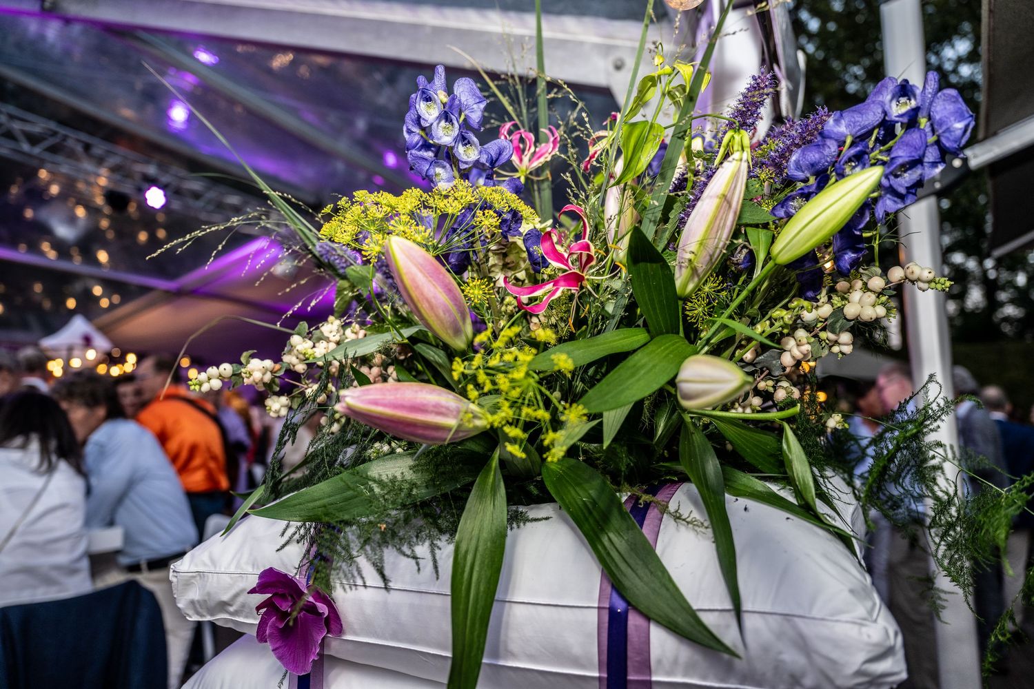 Vibrant floral arrangement with purple iris, pink lilies and greenery displayed at an indoor event venue.