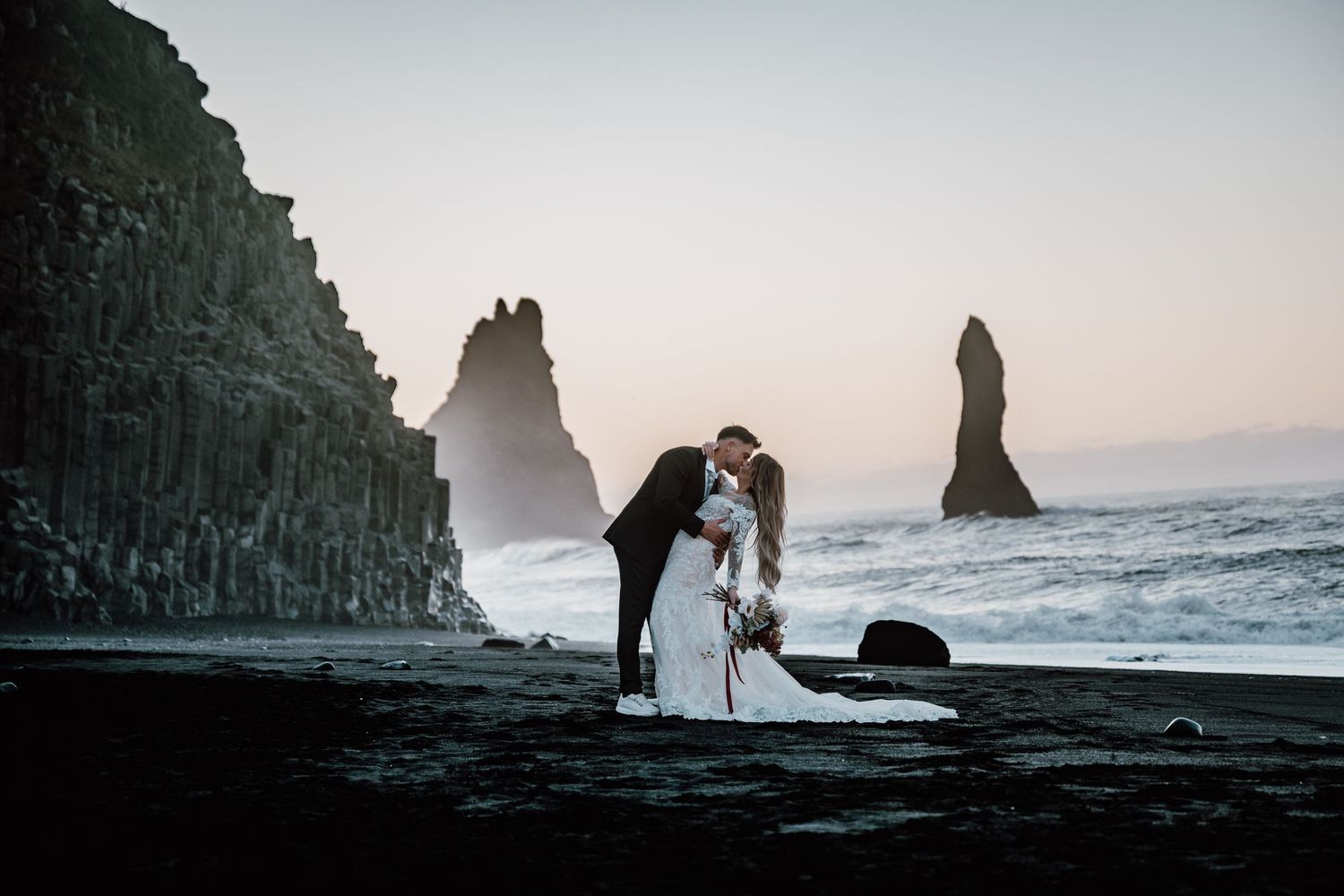 Silhouettes of figures on a dramatic black sand beach with towering rock formations and crashing waves at sunset.