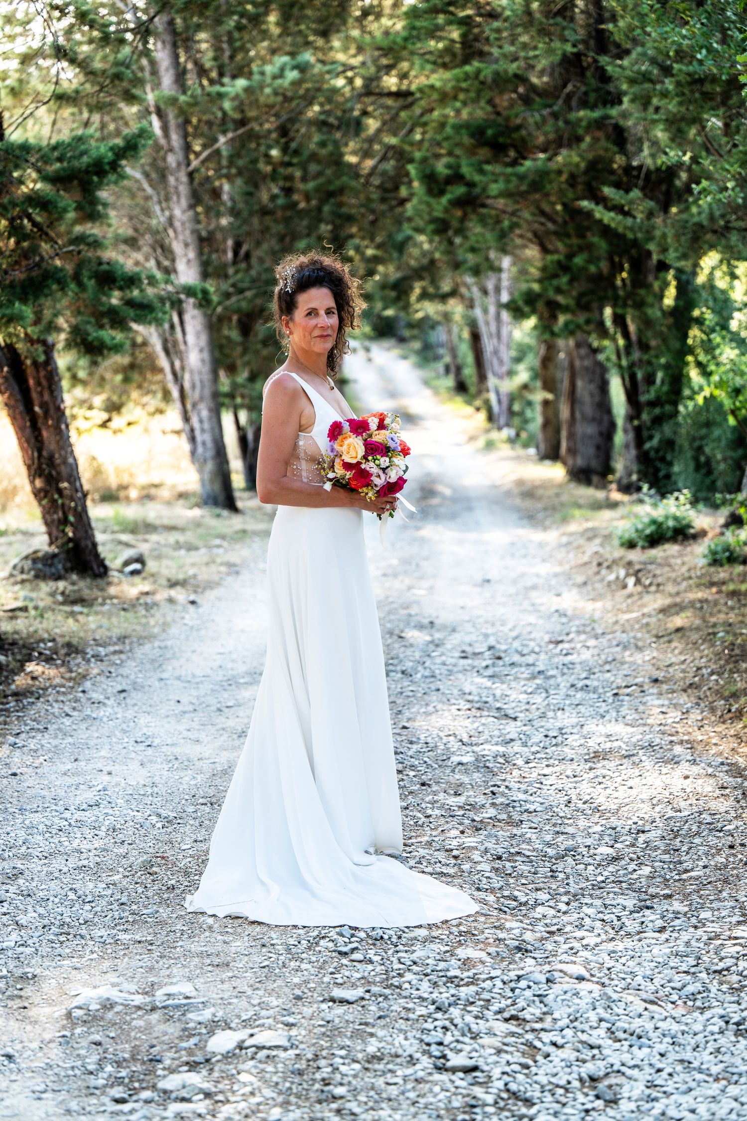 Elegant bride in white gown holds bouquet on forest path.