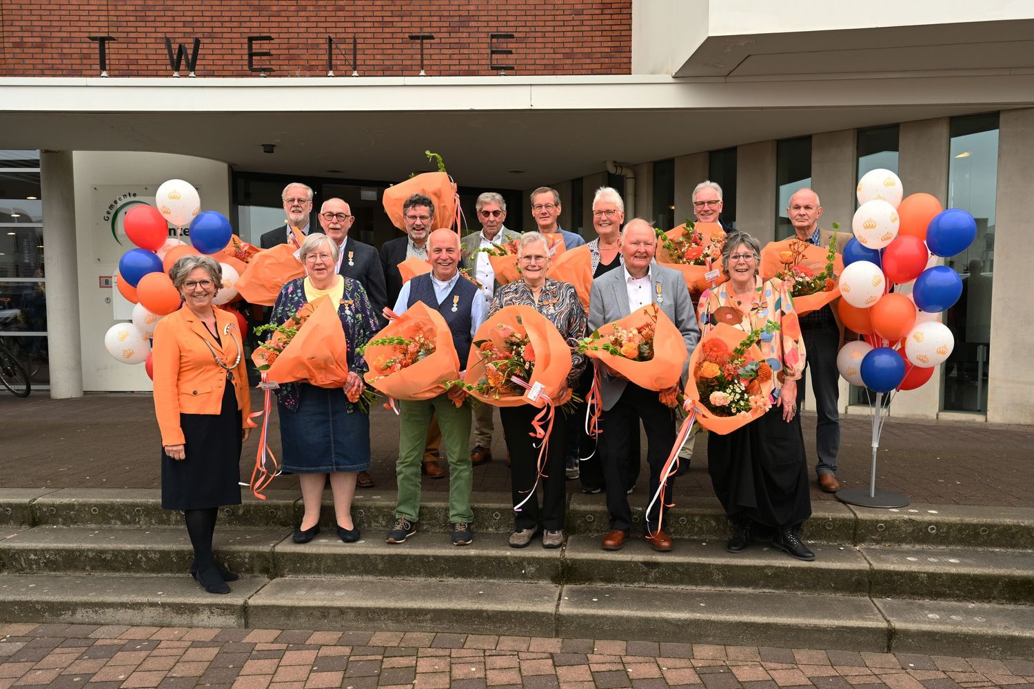 Large group gathering outside a building with orange and blue balloons for a celebration.