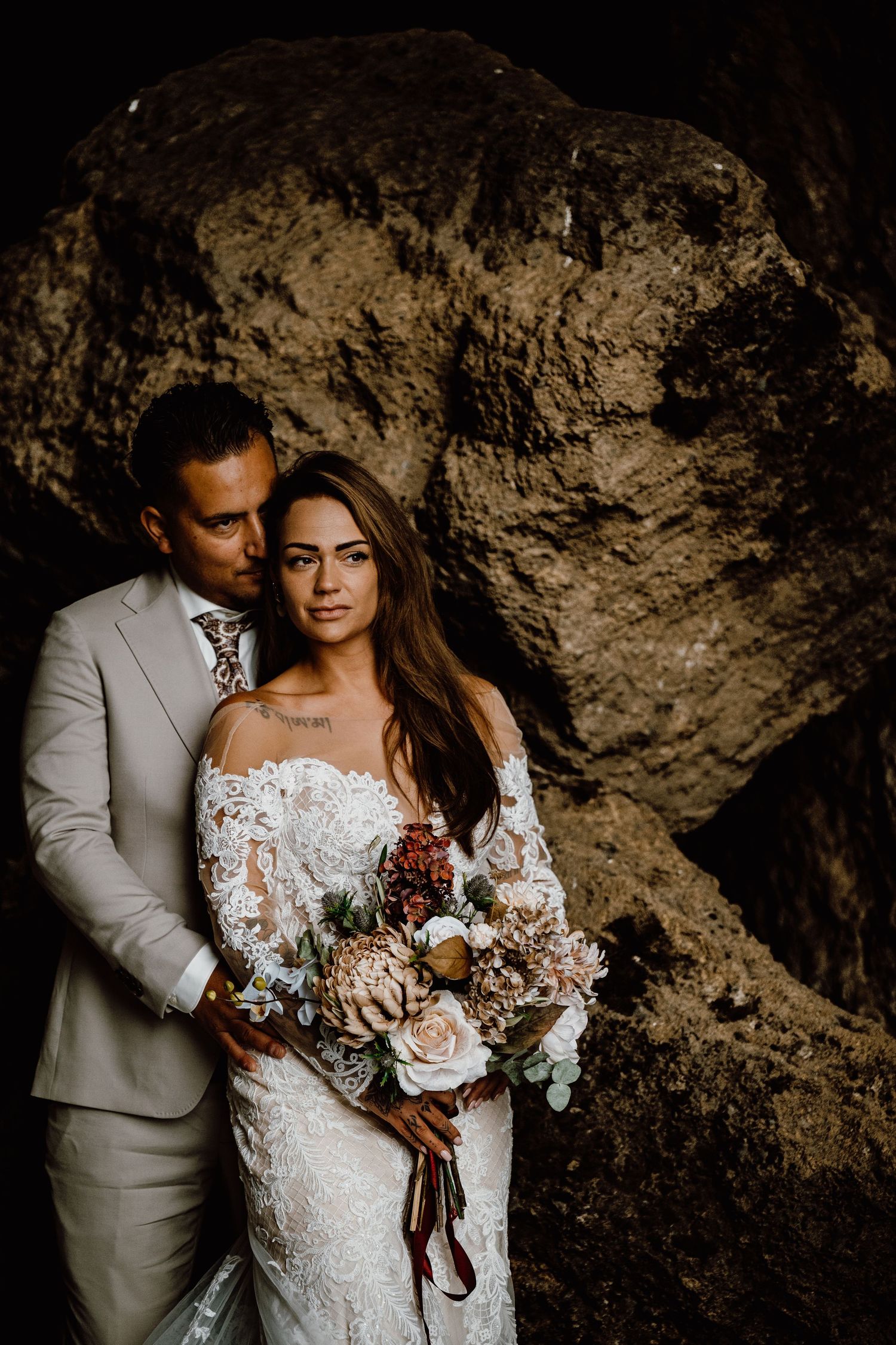 A romantic wedding photoshoot against a dramatic rock wall backdrop showcasing elegant bridal attire and a beautiful bouquet.