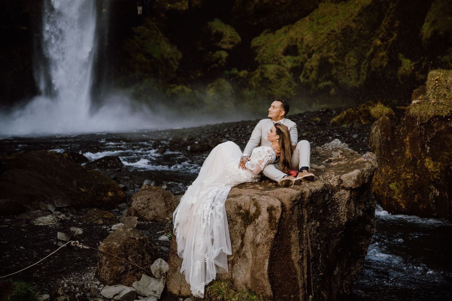 A couple in wedding attire poses dramatically on rocky cliffs near a rushing waterfall.