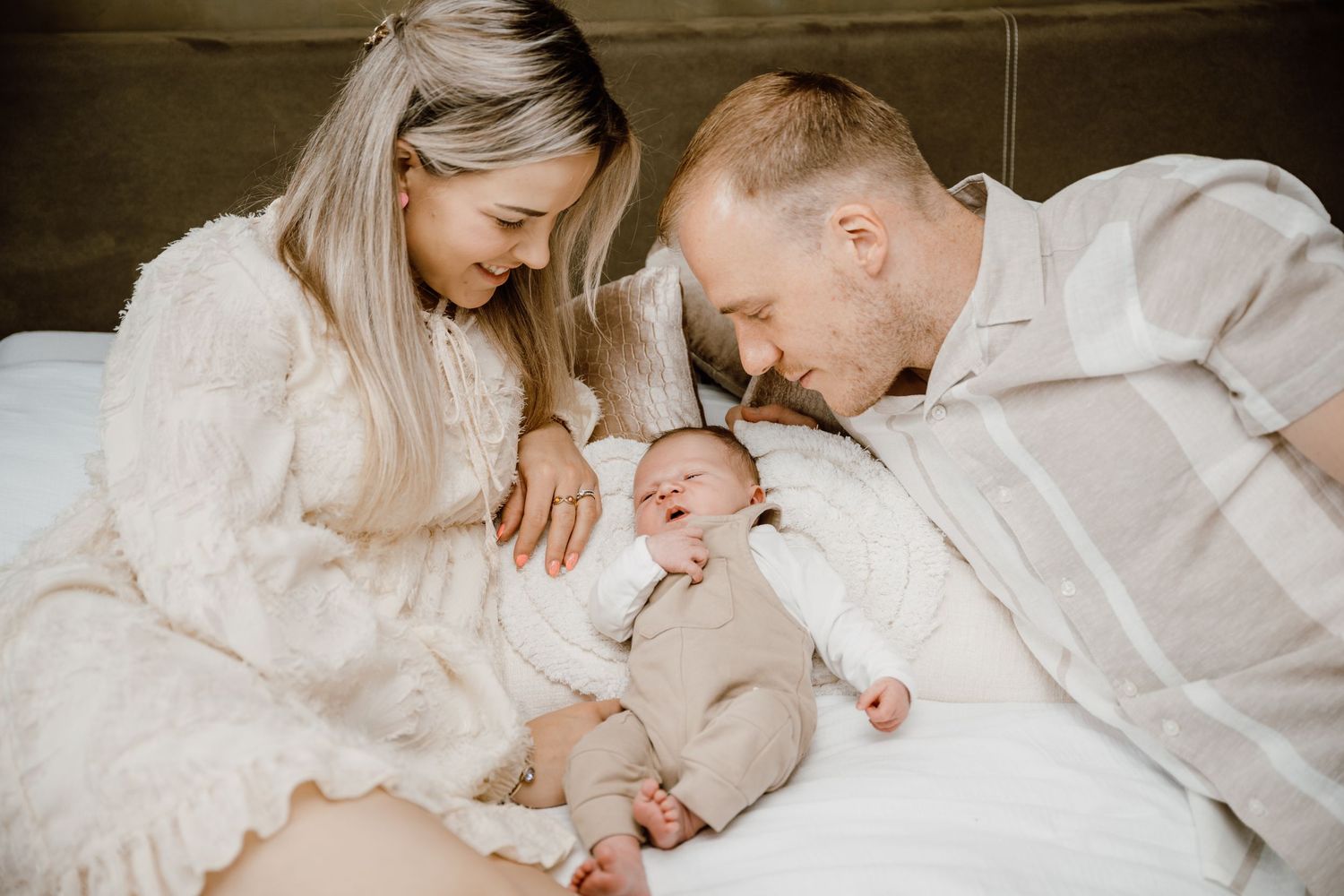 A loving family shares tender moments together while lying on white bedding in a bright, airy setting.