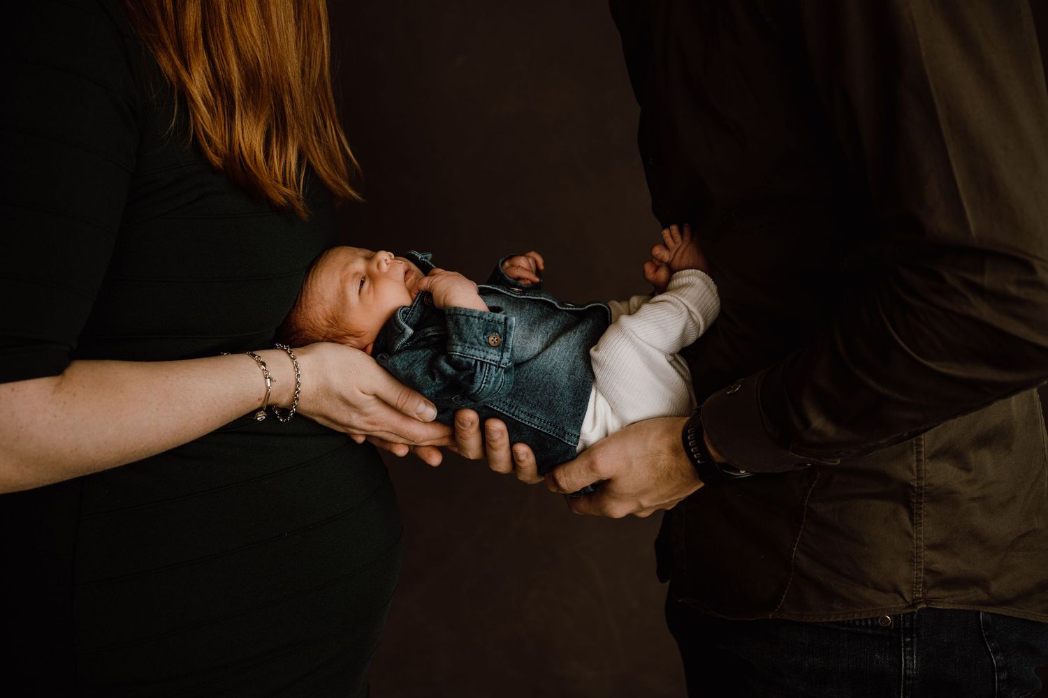 Two people holding a newborn baby wrapped in a gray blanket against a dark background.