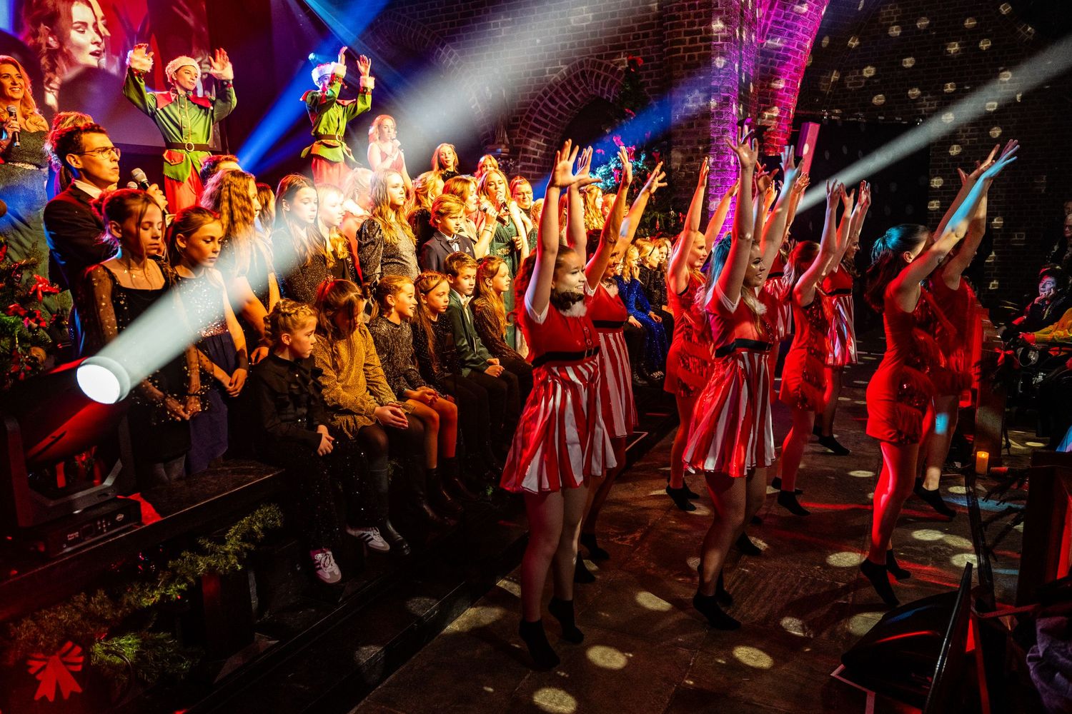 Large group of performers on stage dressed in red costumes under vibrant stage lighting.