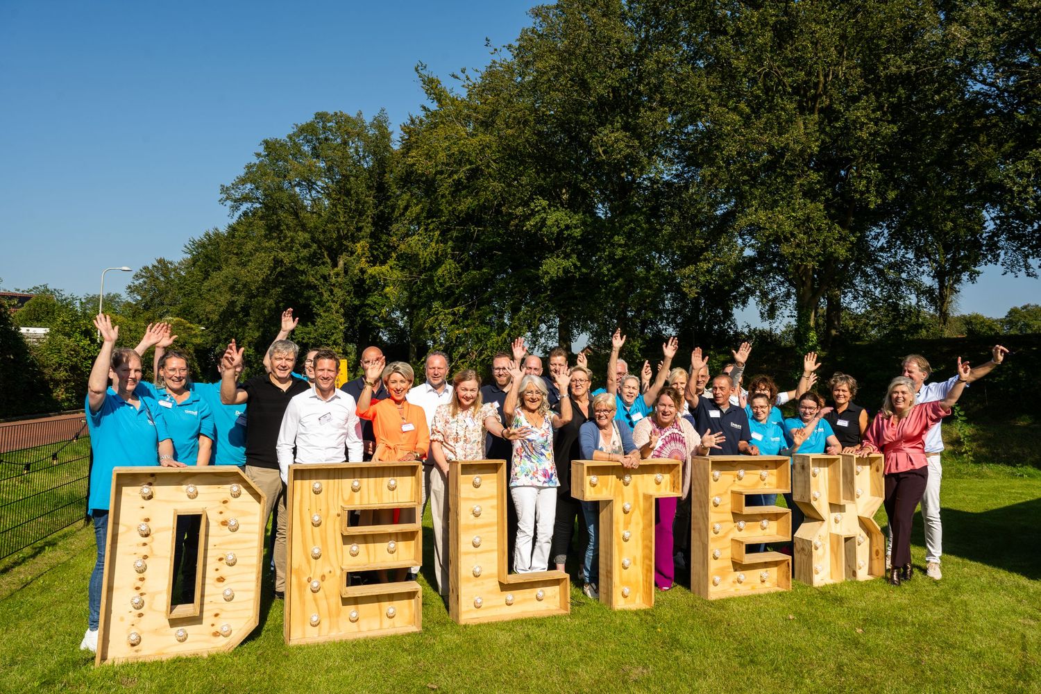 Large group of wedding guests pose behind giant wooden DELITES letters on a sunny grassy hillside.