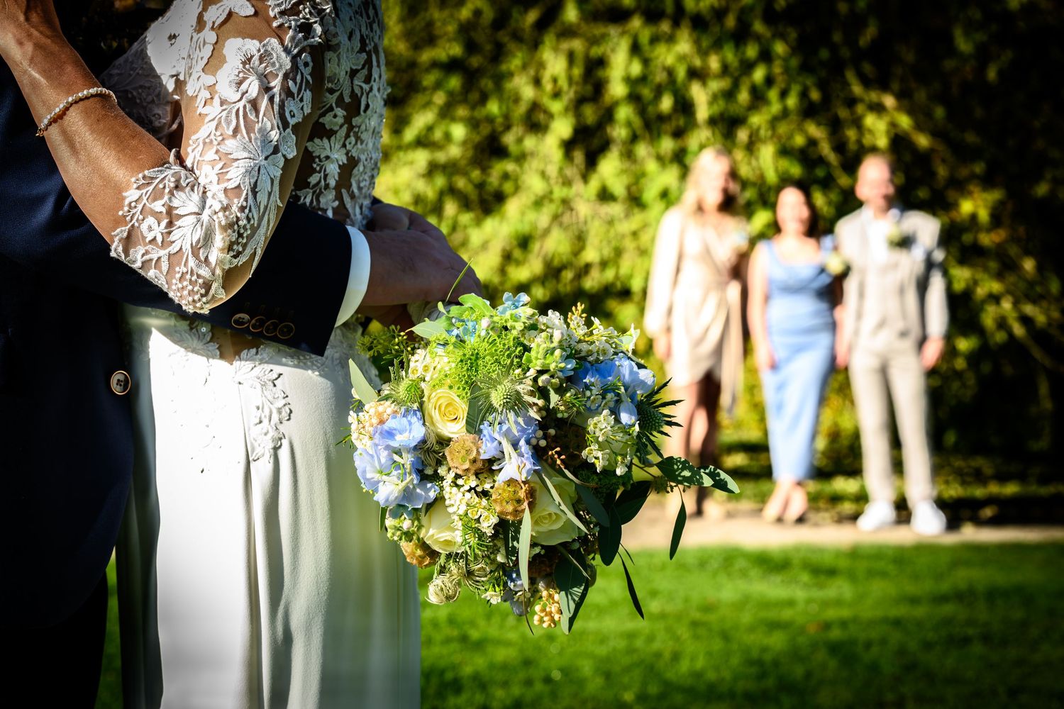 A couple poses in a garden setting wearing formal attire with sunlit trees in the background.