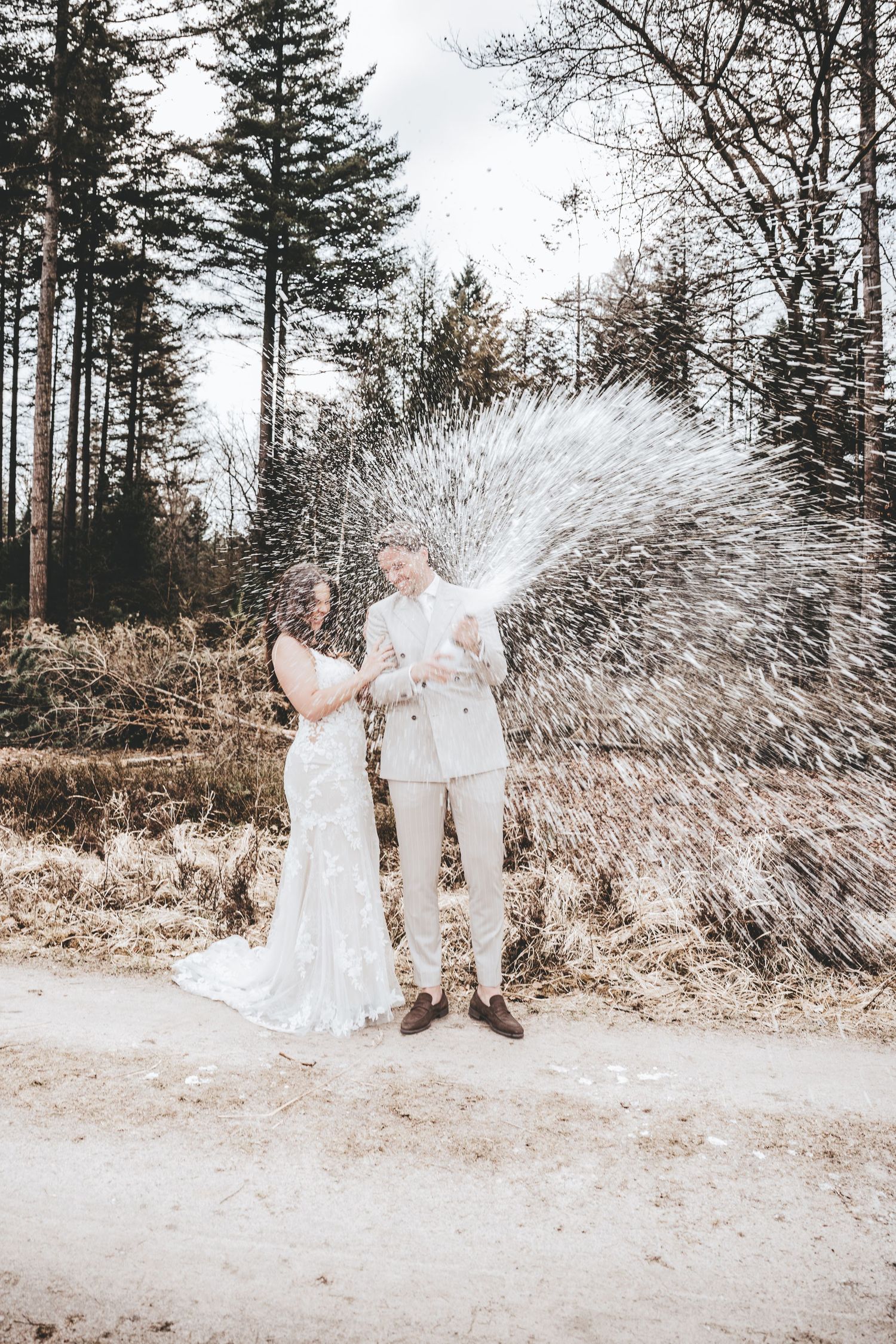 A couple in white attire popping champagne in a forest setting during a celebration.