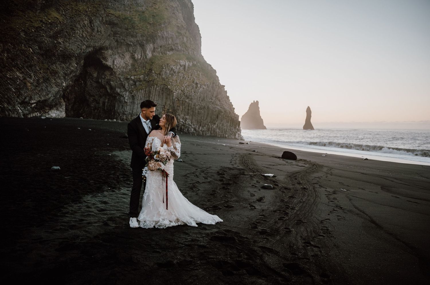 A romantic beach wedding photoshoot featuring a couple against dramatic black sand and coastal rock formations at sunset.