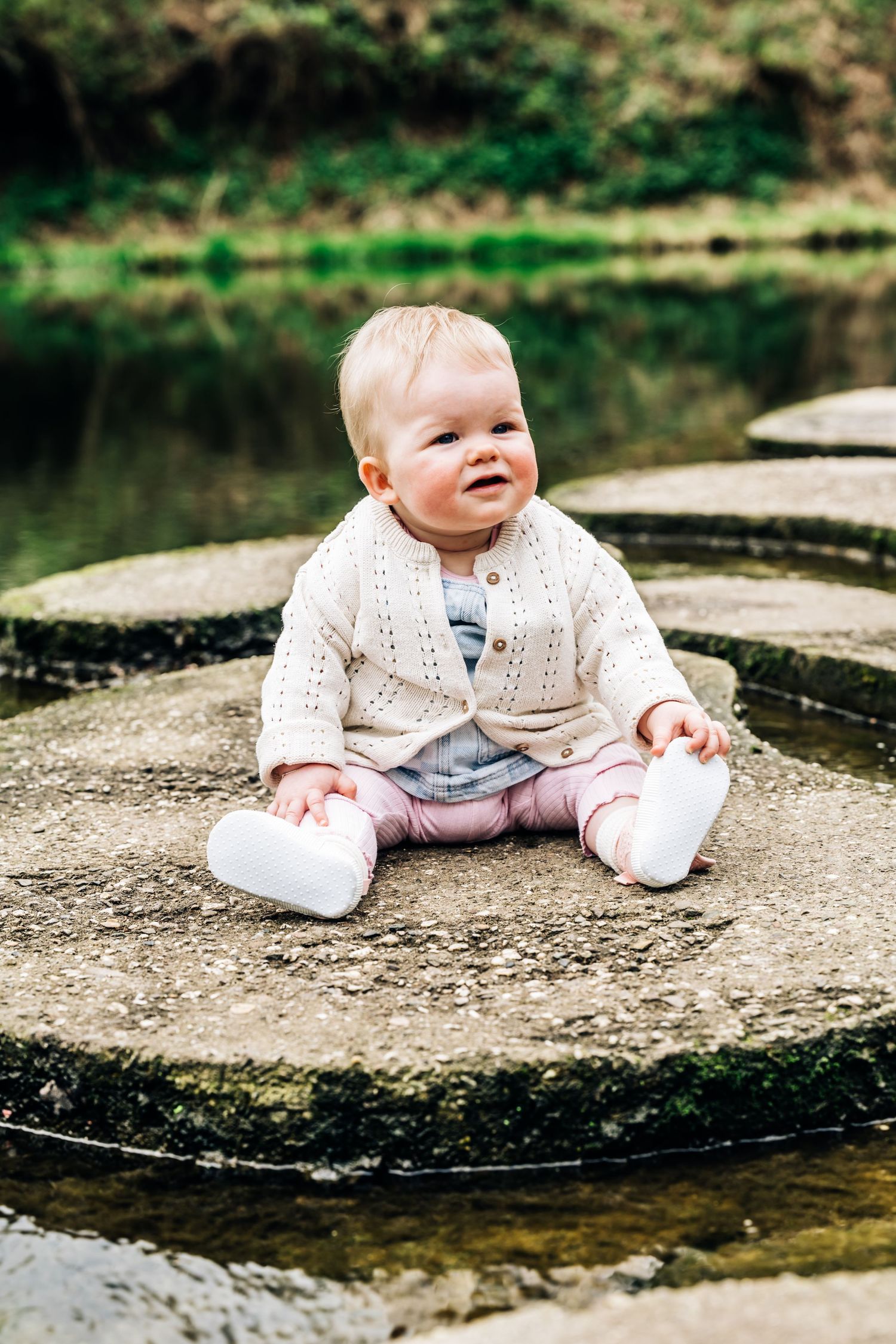 Baby in white knit sweater sitting on circular stone platform near water.