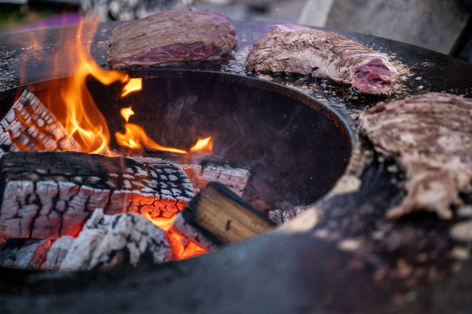 Thick steaks grilling over hot flames and glowing embers on a cast iron grate.