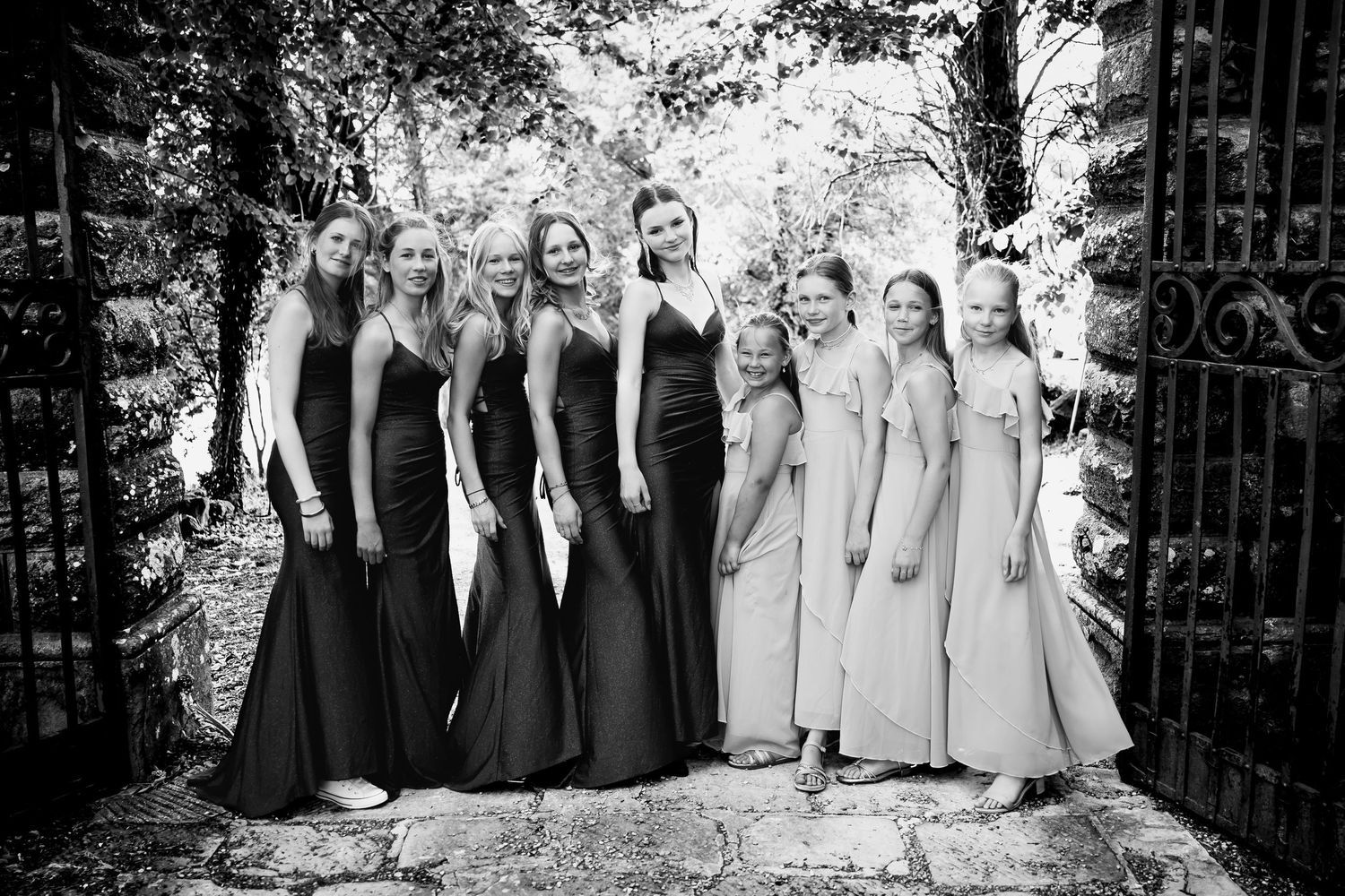 Group photo of bridesmaids in formal dresses standing by garden gate.