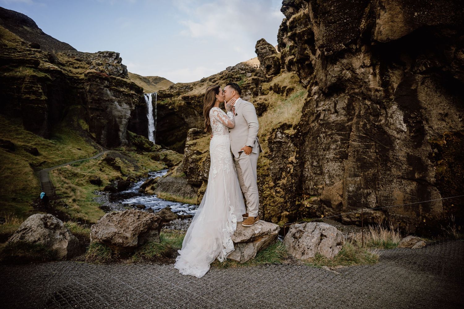Couple in wedding attire embraces near dramatic rocky cliffs and waterfalls in Iceland's rugged landscape.