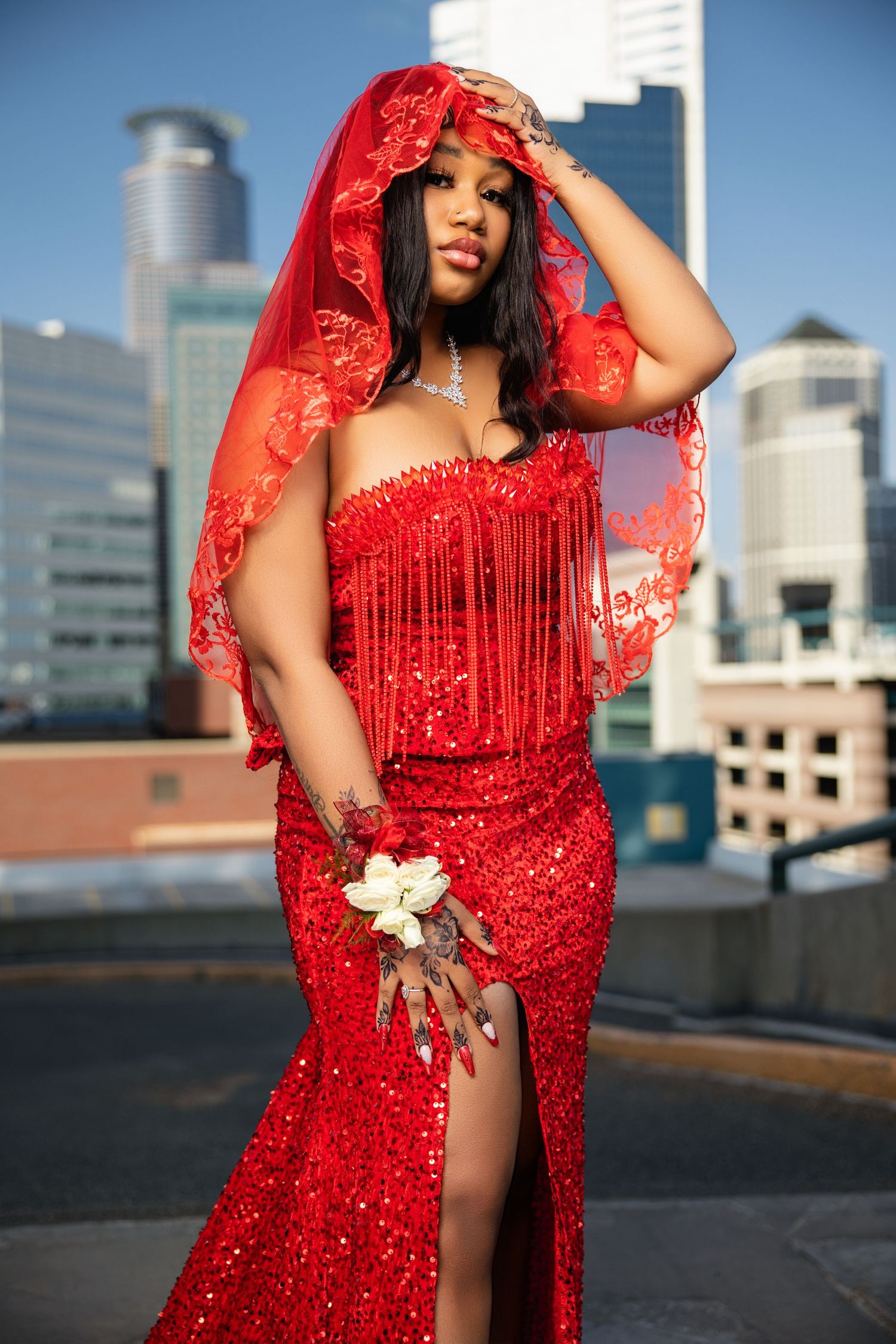 Prom outdoor portrait. Model wearing red dress with red head covering. Posing with one hand placed on head, and the other on thigh.