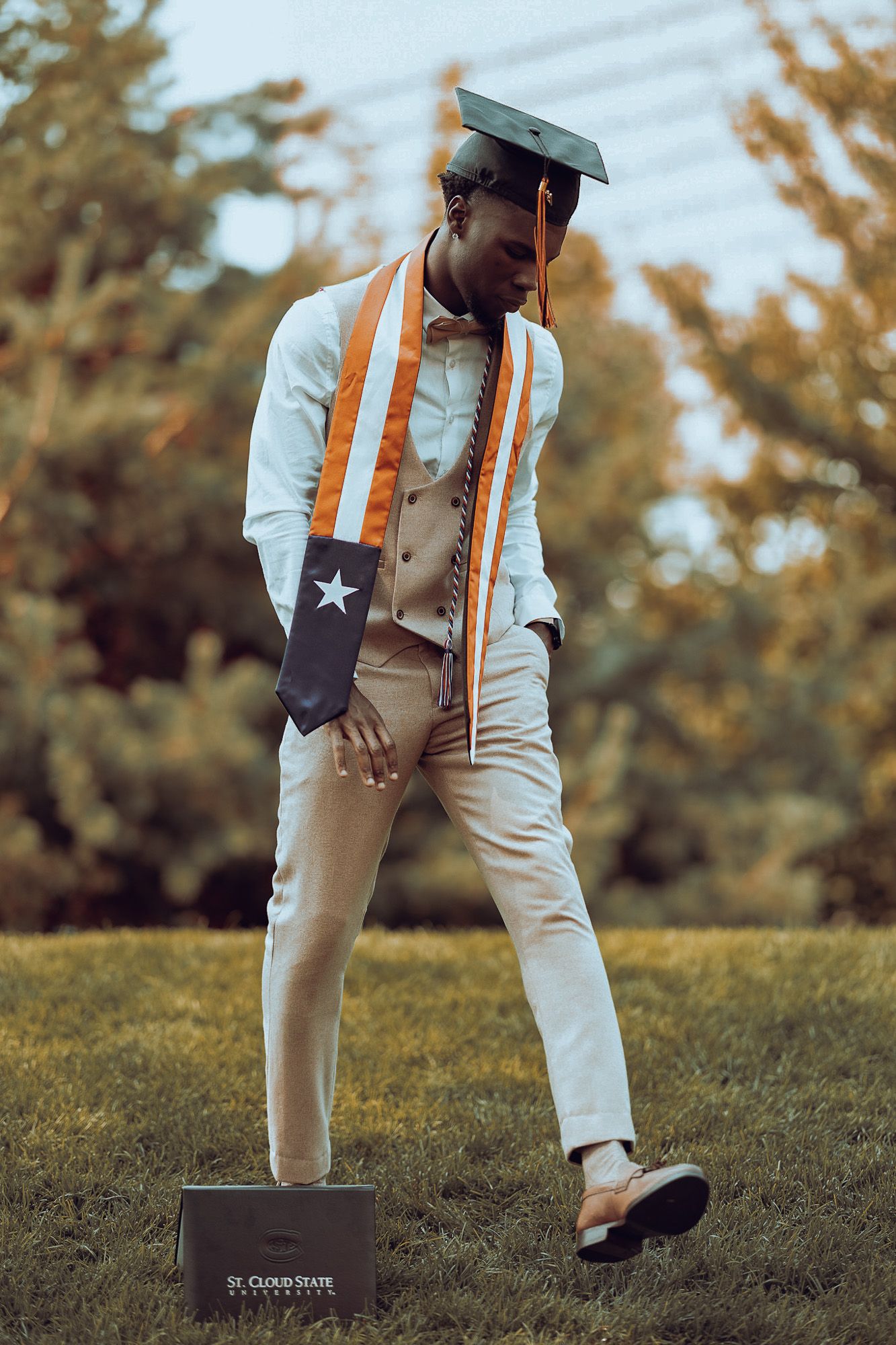 Graduation Portrait. Model is wearing his grad cap, grad (Liberian flag) sash around neck, looking down, and kicking one leg slightly up.