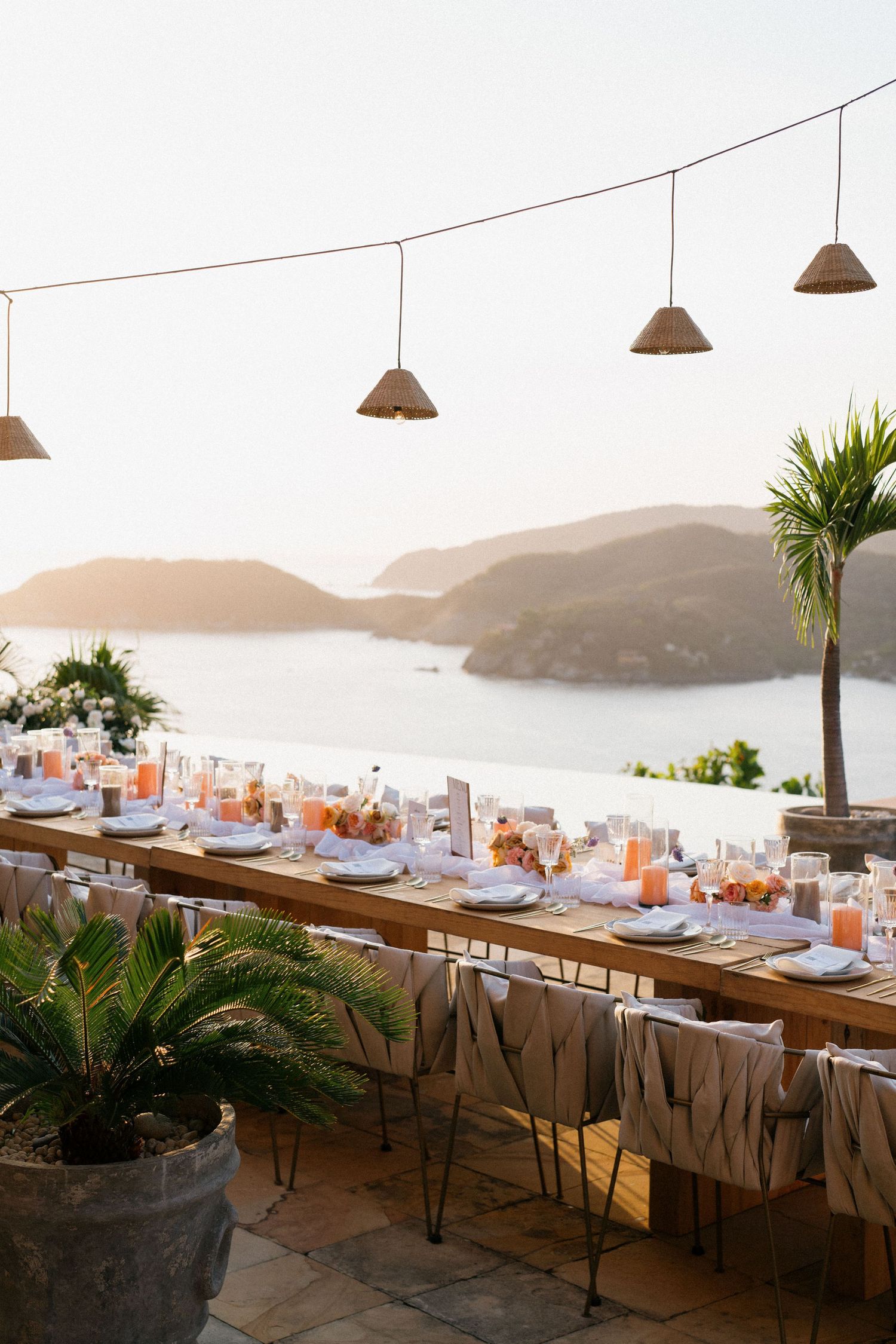 An elegant outdoor wedding reception setup with long tables overlooking a coastal bay view at sunset.
