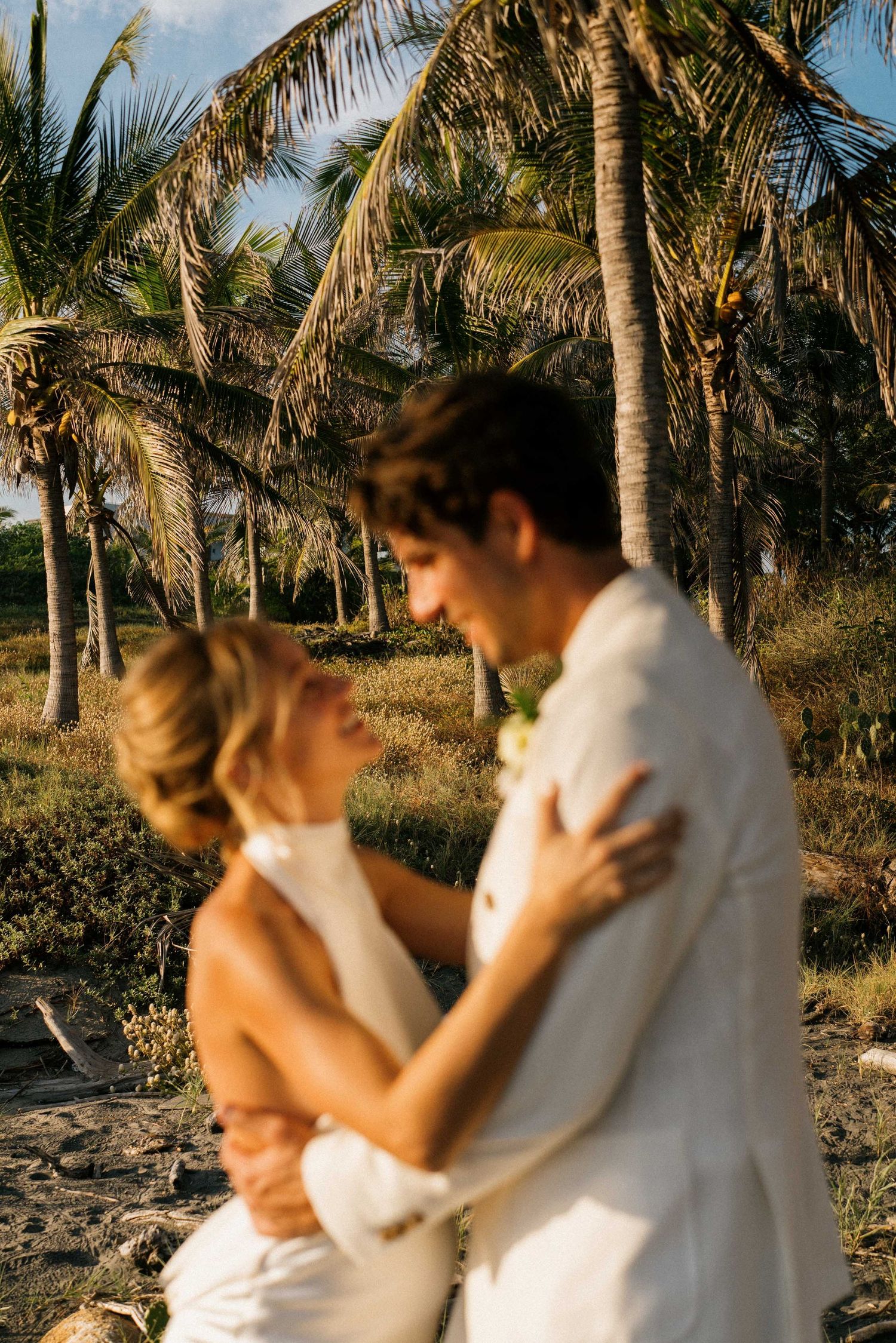 Intimate beach photoshoot with couple in white attire against a backdrop of palm trees at sunset.