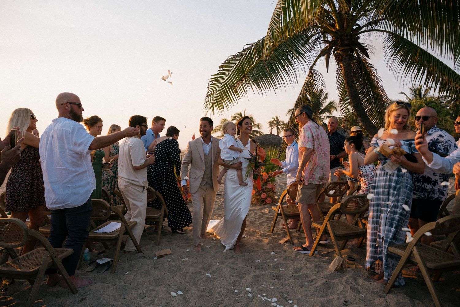 Wedding guests gathered on wooden chairs at a palm-fringed beach ceremony.