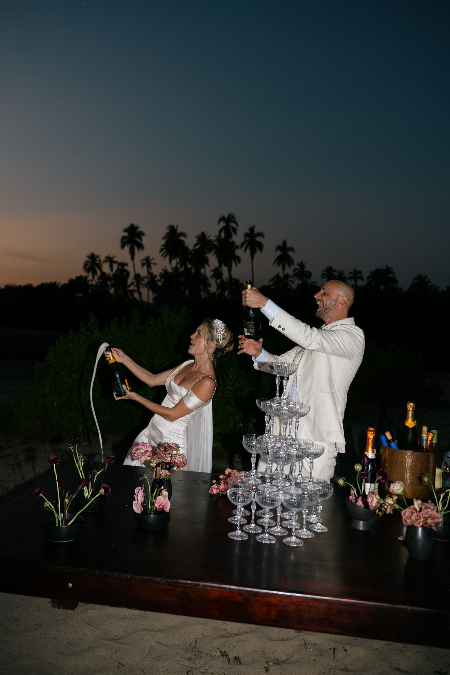 Couple pouring champagne into tower of glasses at sunset with palm trees in background.