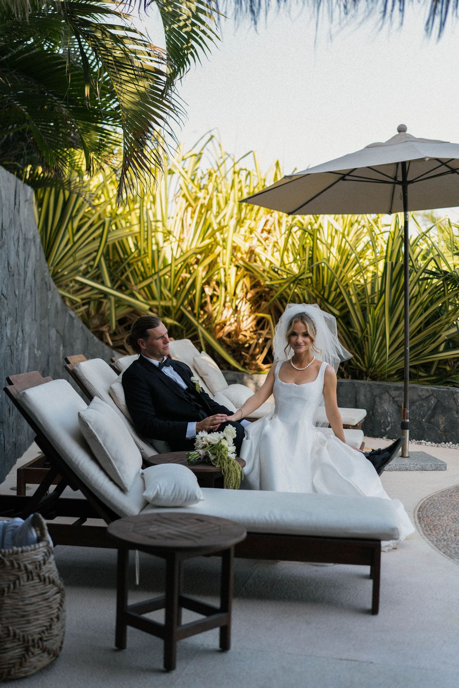 Relaxed wedding couple lounging on patio chairs surrounded by tropical plants.