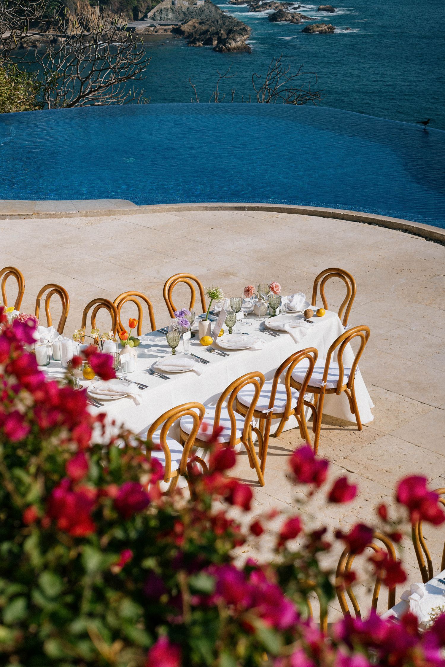 An elegant outdoor wedding table setting with ocean views, wooden chairs and pink bougainvillea flowers.