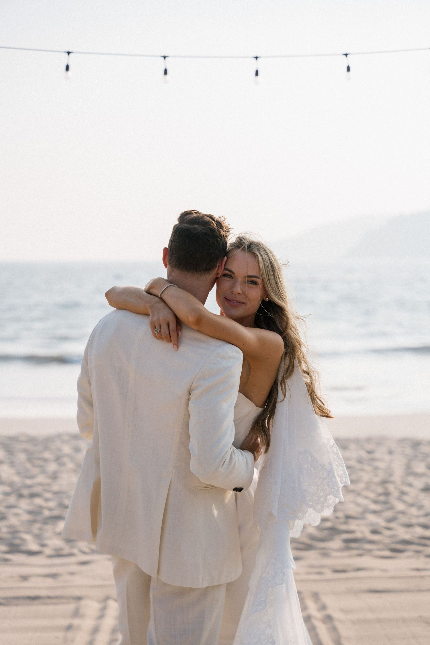 Romantic beach embrace between couple wearing all white against ocean backdrop.