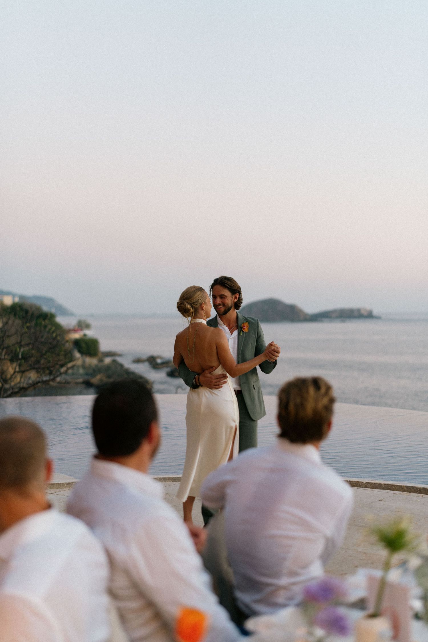 Seaside wedding ceremony with guests in white attire overlooking the ocean at sunset.