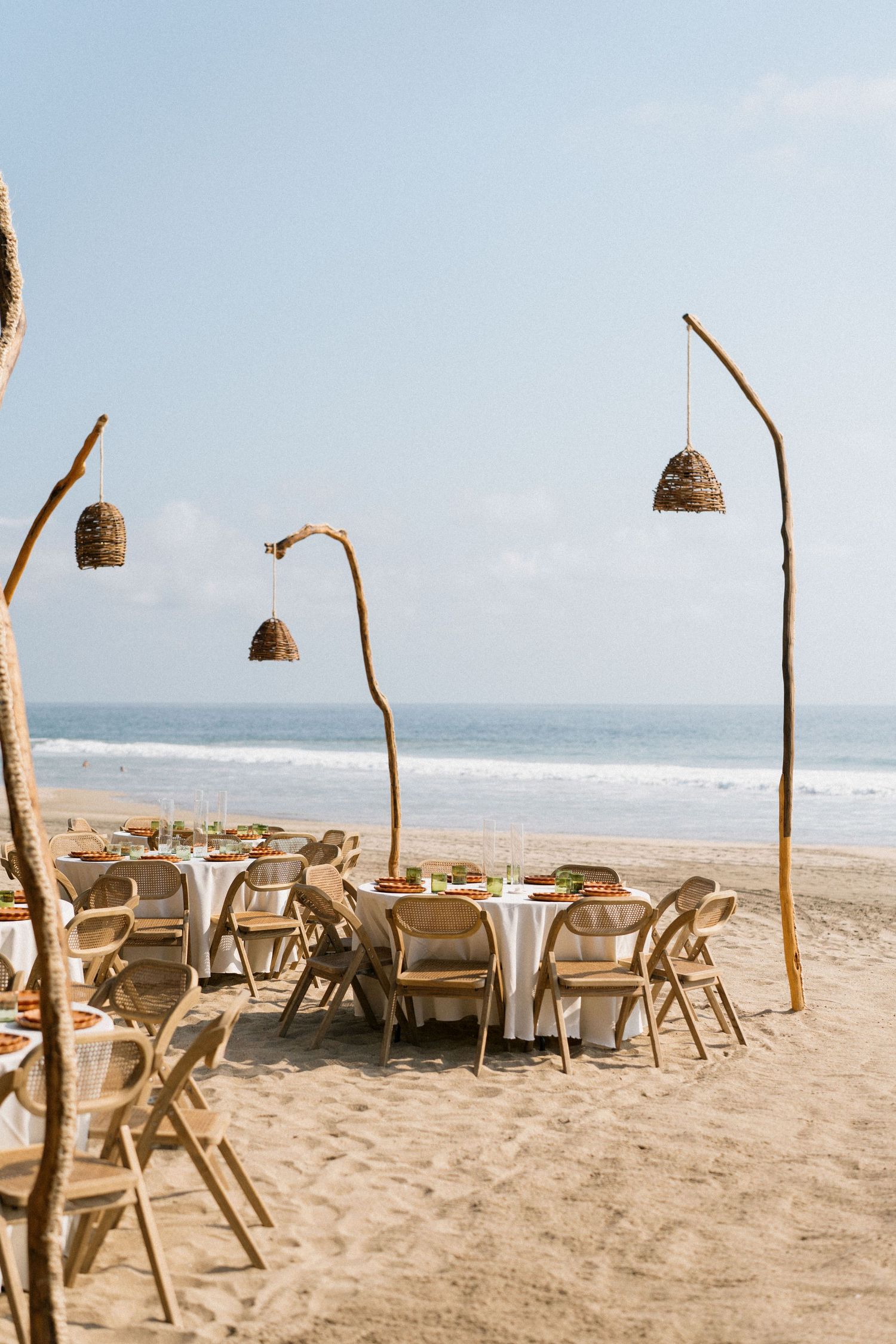 Beach wedding setup with rattan pendant lights and natural wooden chairs on sandy shore.