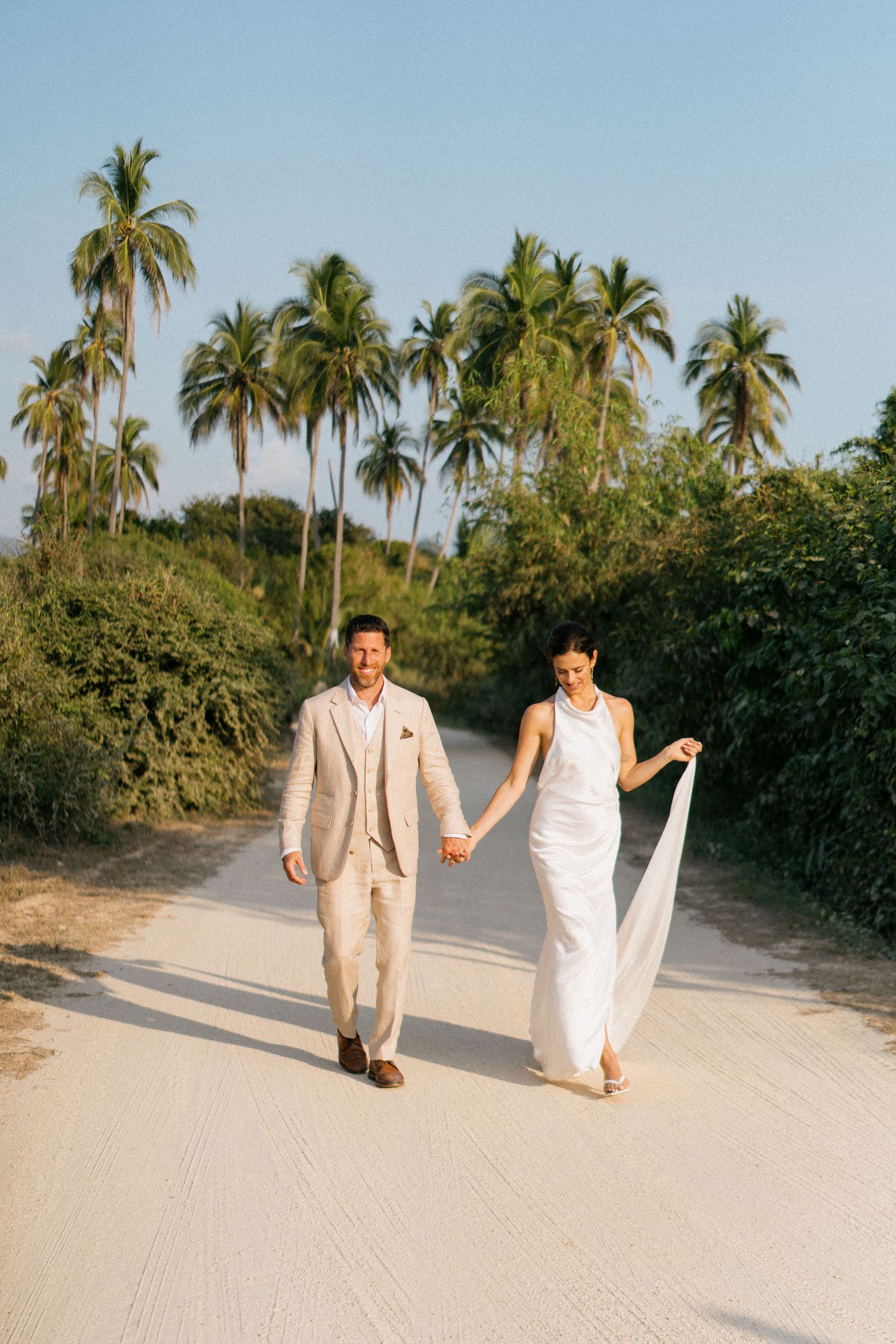 A couple walks hand in hand along a sandy beach path lined with swaying palm trees in tropical paradise.