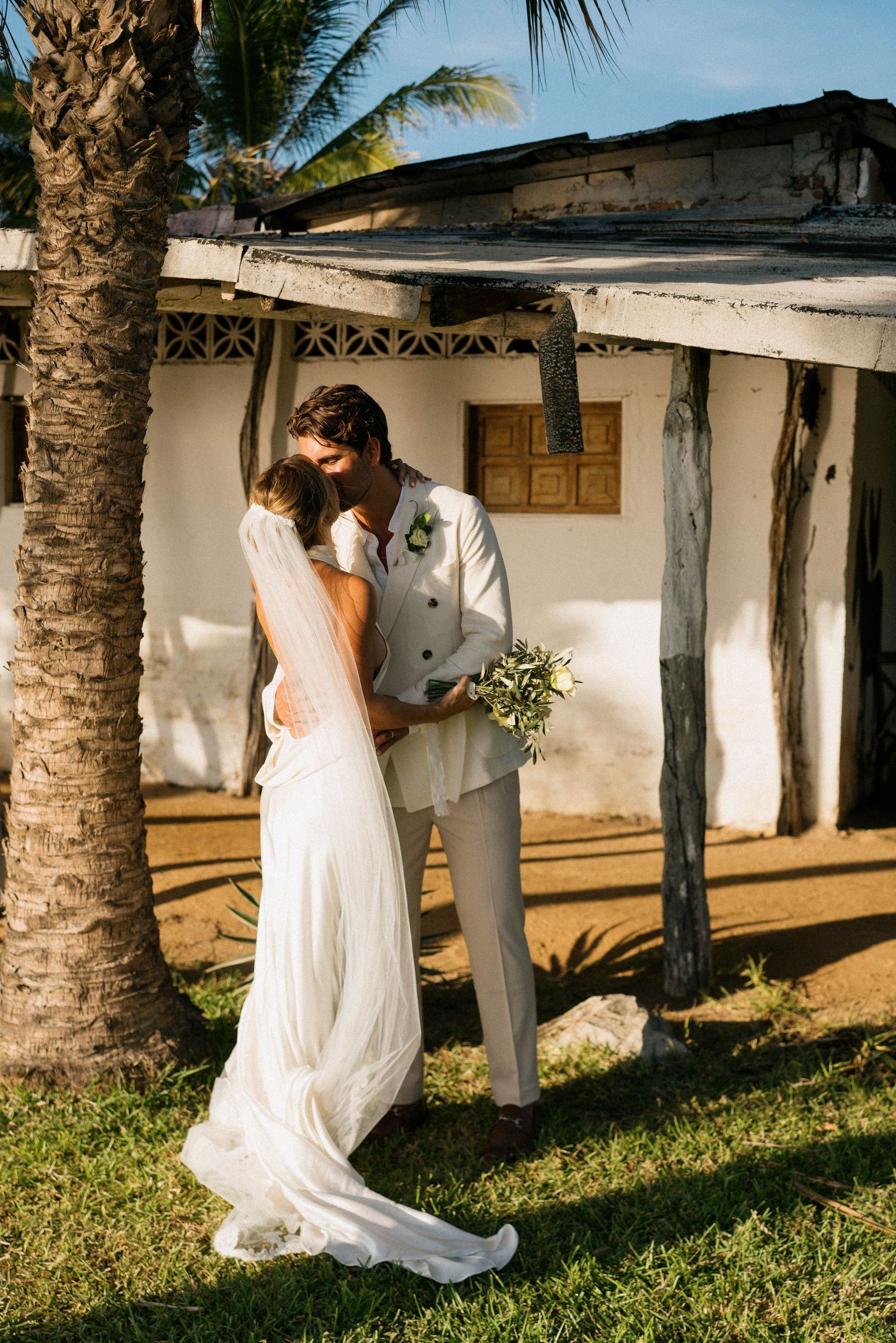 A romantic wedding moment under a palm tree by a rustic white building.