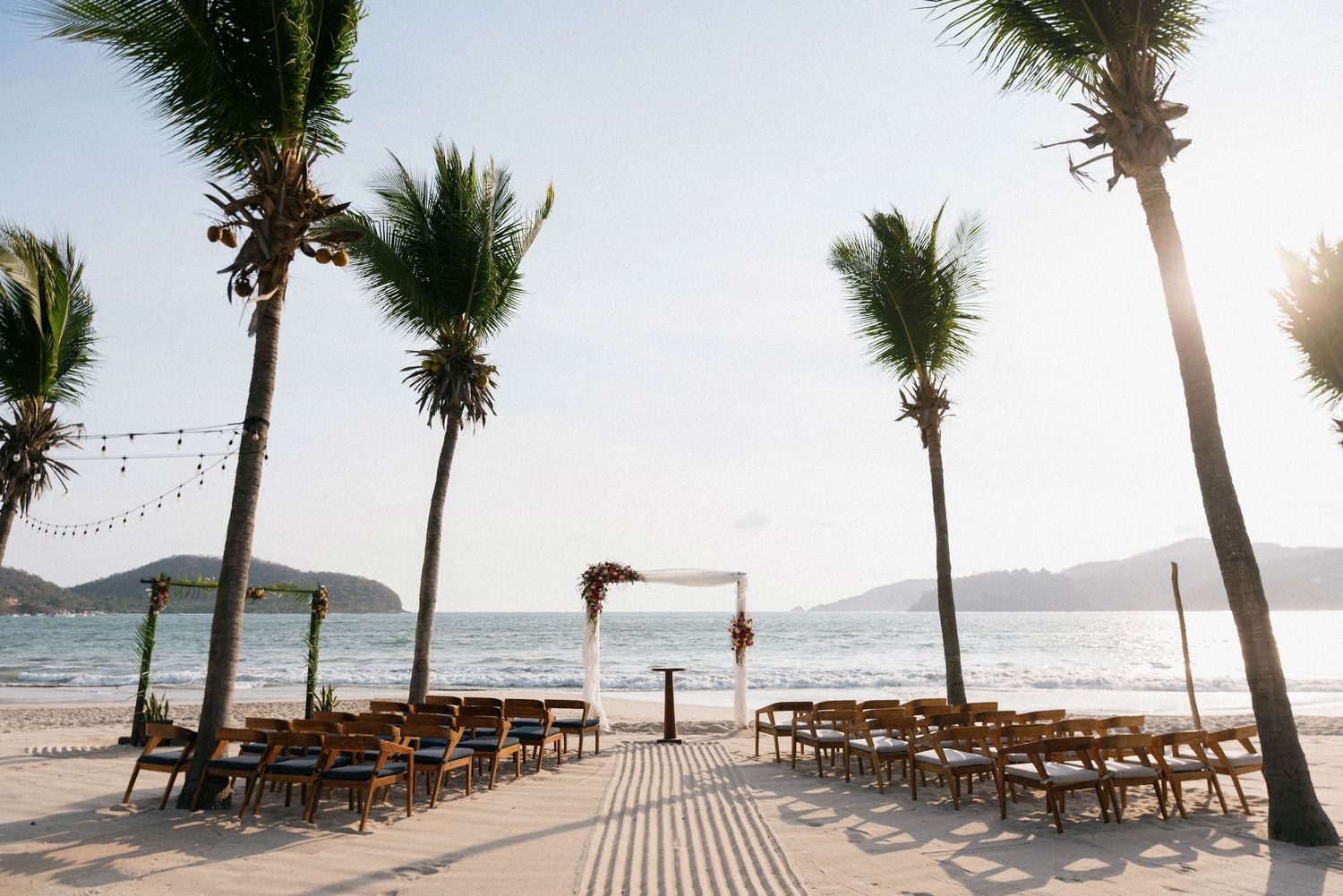 Empty beach ceremony setup with wooden chairs and palm trees along tropical shoreline.