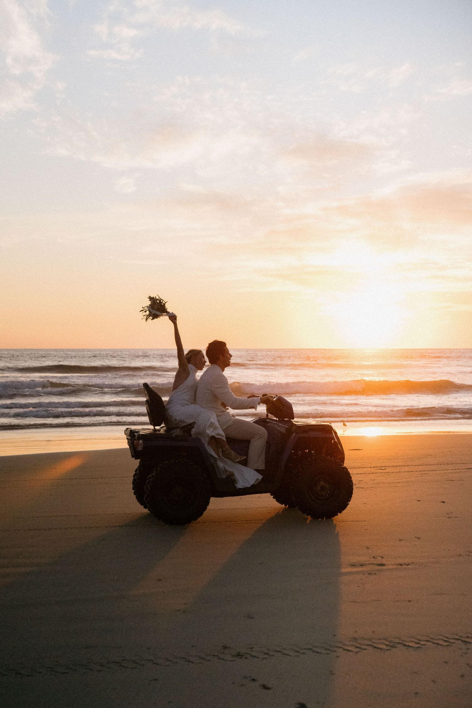Newlyweds riding an ATV on the beach at sunset holding a bouquet.