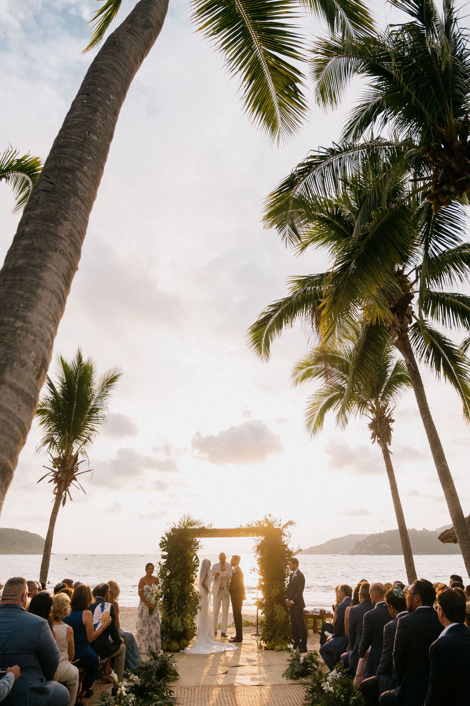 Beach wedding ceremony under palm trees with guests seated facing an ocean view.