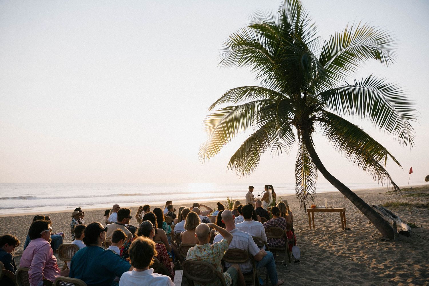 Beachfront wedding ceremony setup with palm trees and ocean views at sunset.