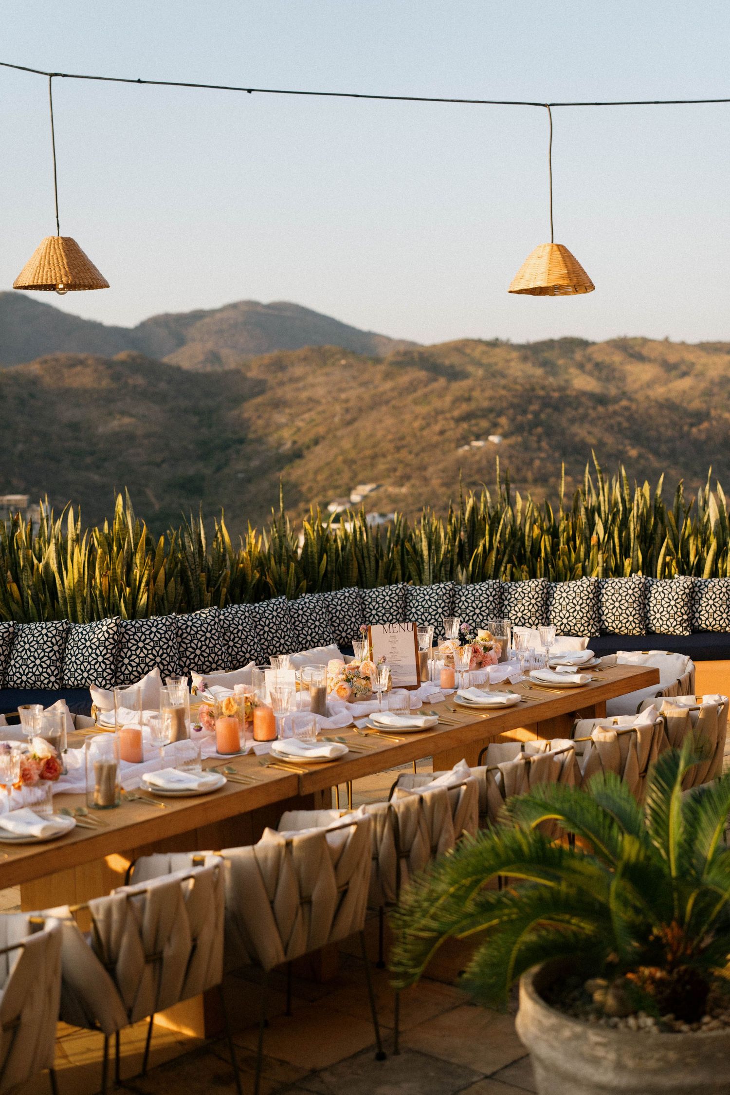Long wooden dining table set up outdoors with mountain views and string lights for a wedding reception.