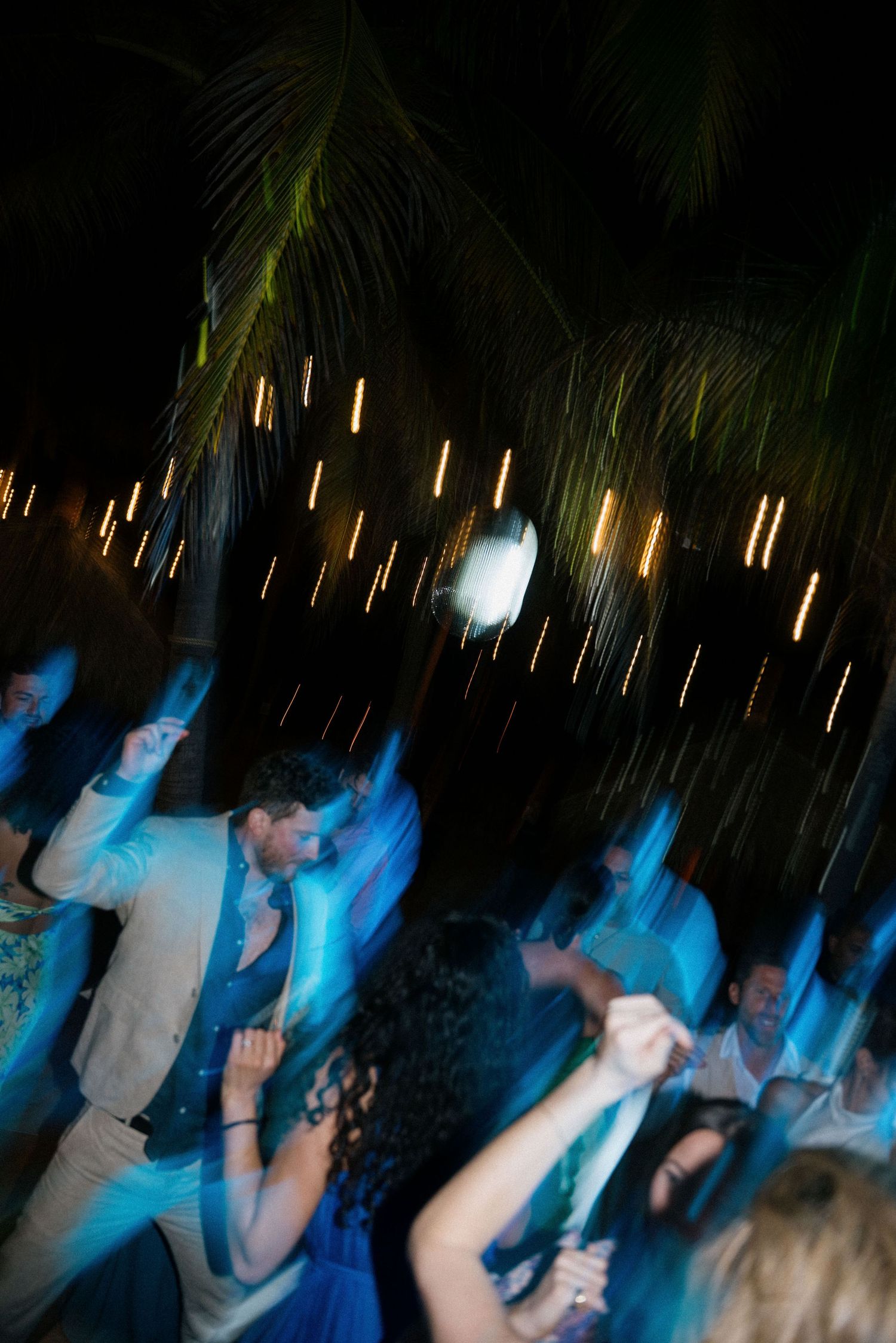 Motion-blur capture of nighttime dancing under hanging lights and palm trees.