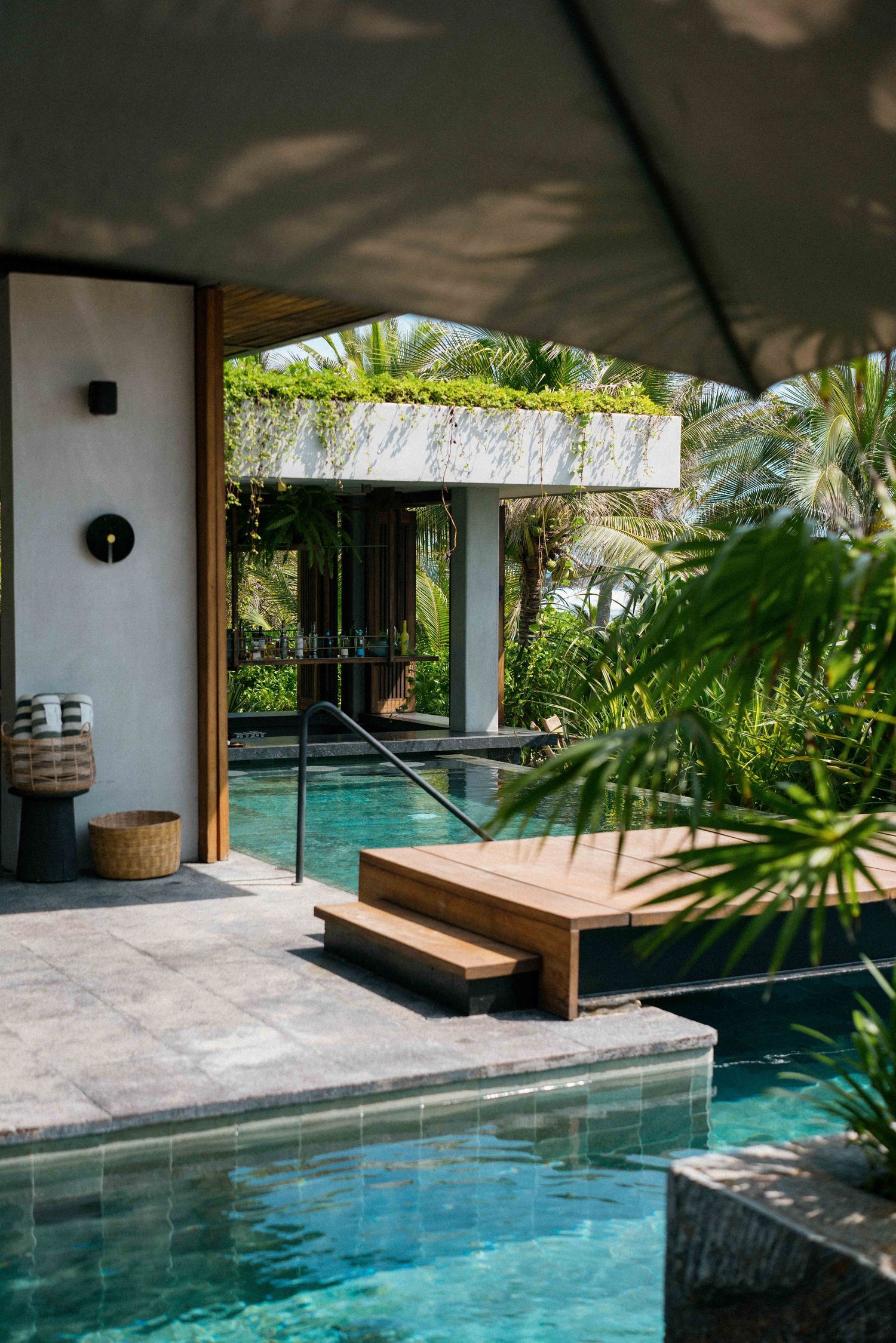 Modern indoor-outdoor swimming pool with wooden deck and tropical landscaping surrounded by white columns and palm trees.