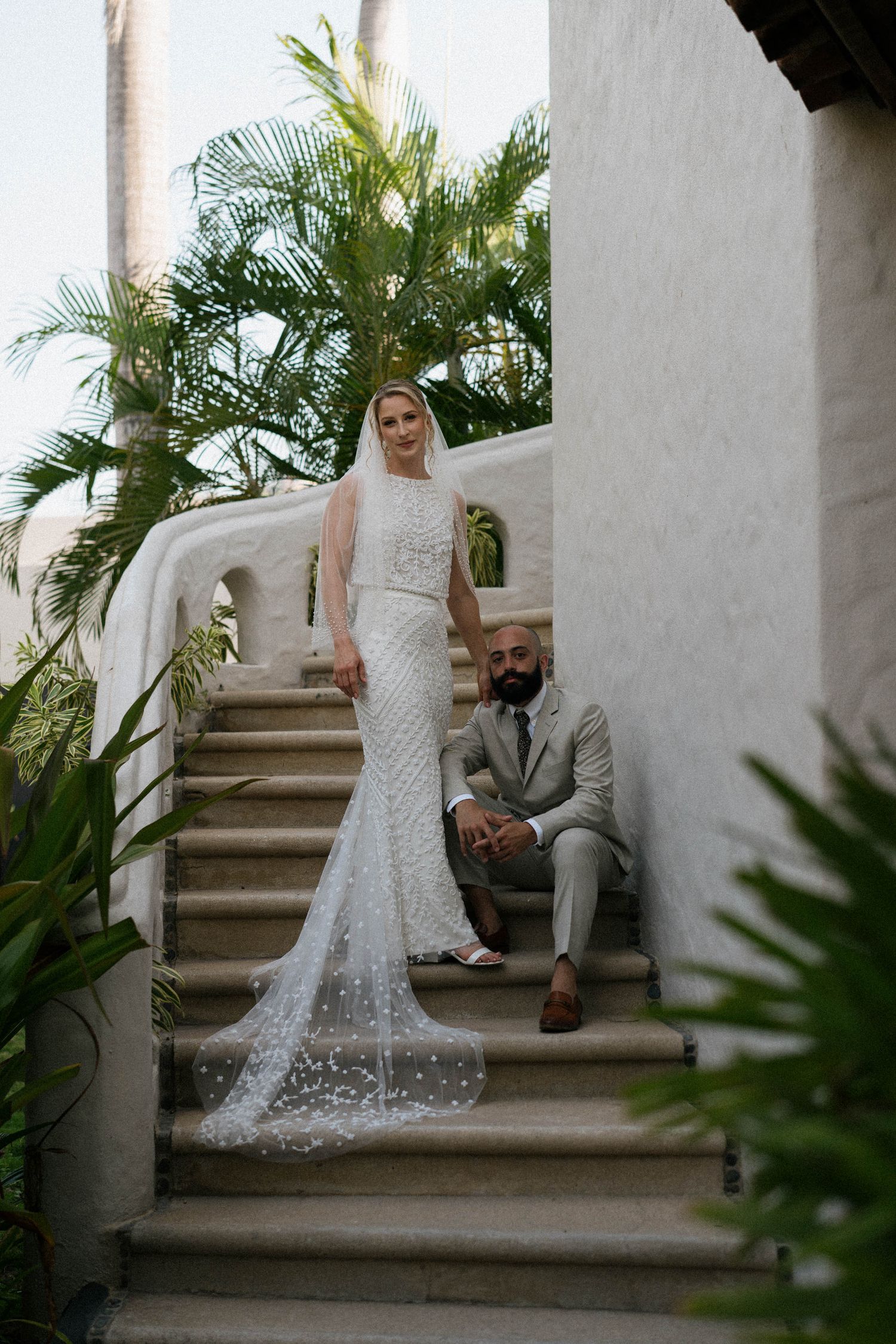 Couple sits together on white stucco stairs surrounded by tropical greenery.