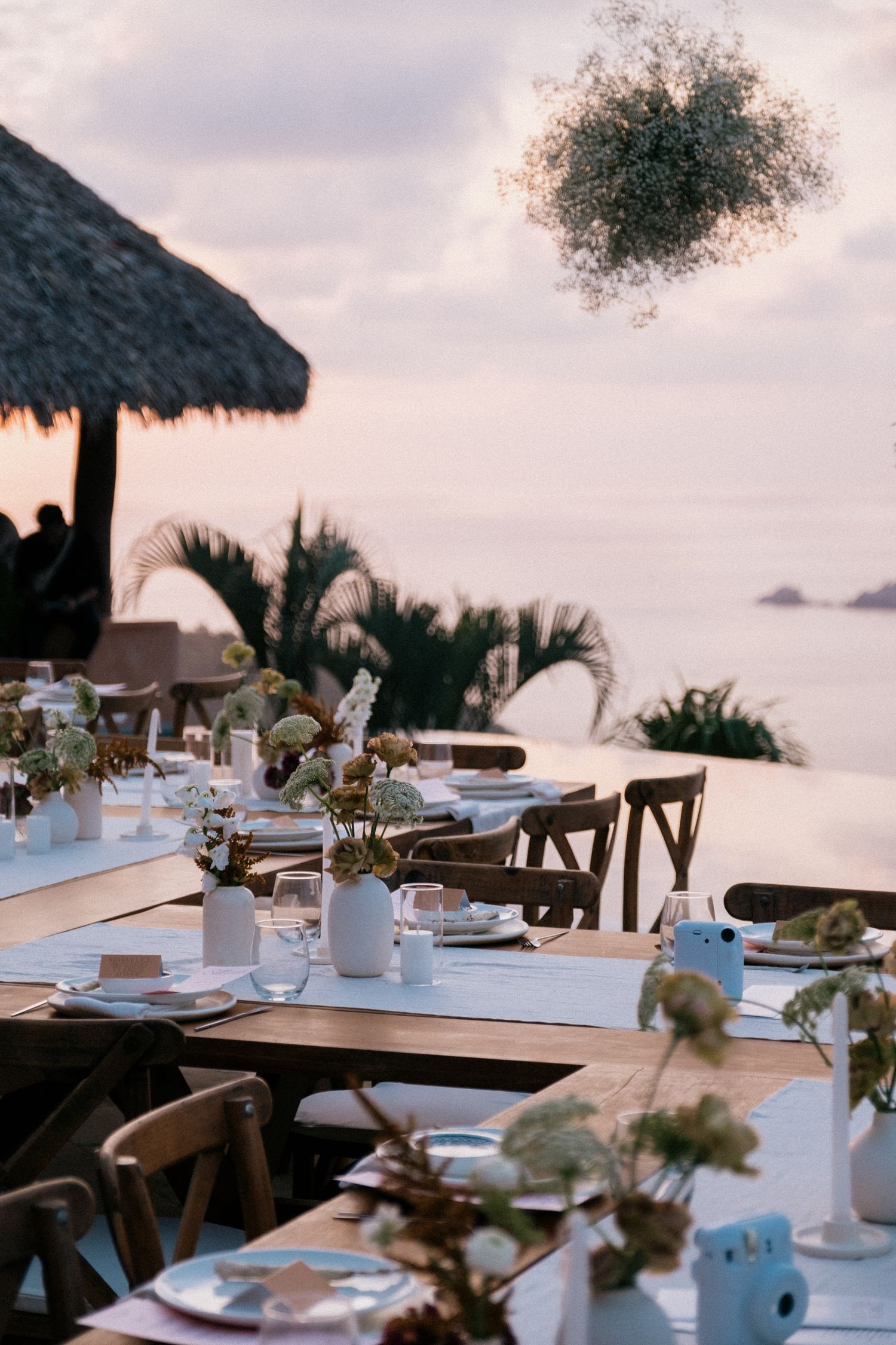Beachfront wedding reception table setup under thatched roof palapa at sunset.