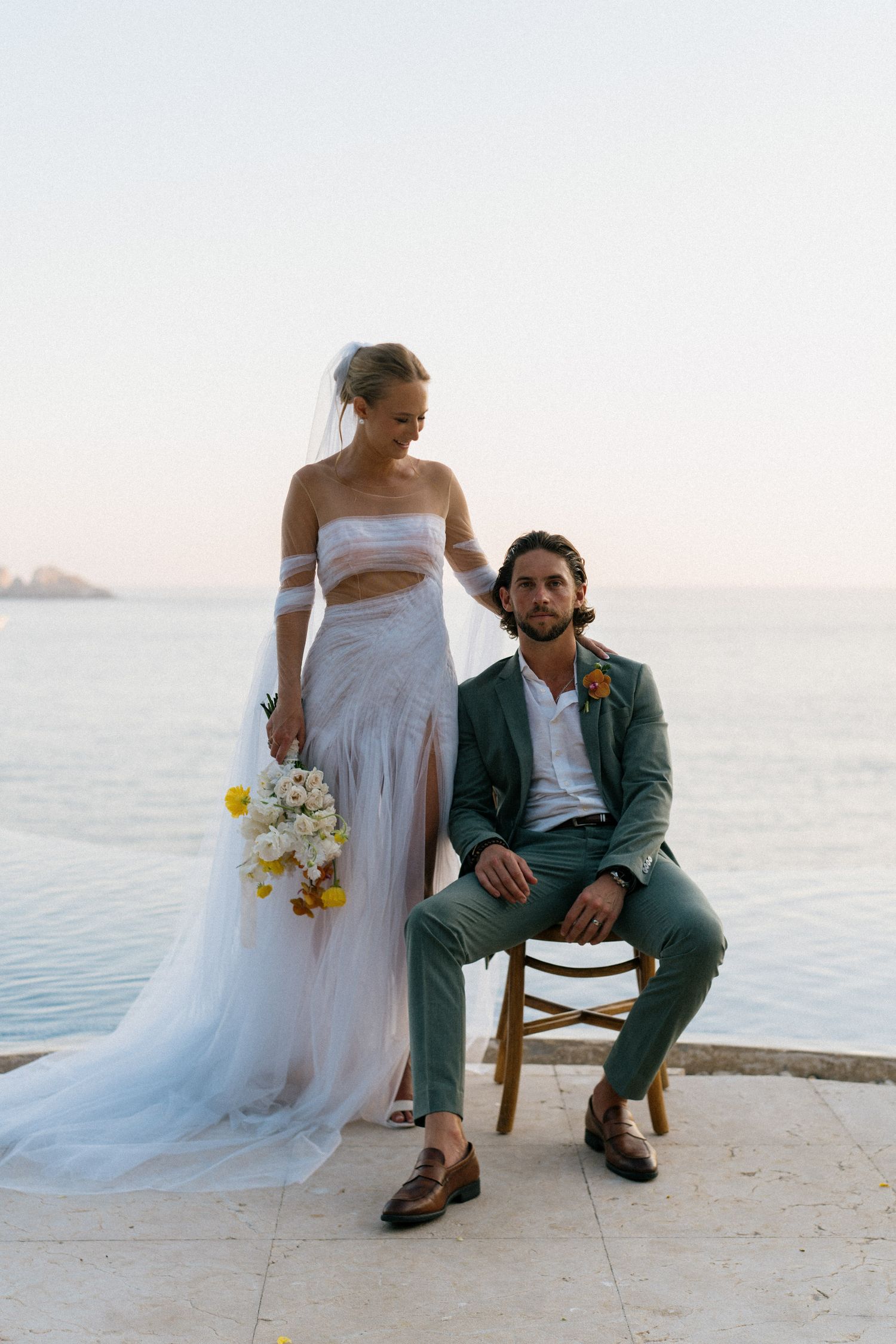 A couple poses for a seaside wedding photo with the bride in white and groom in green suit.