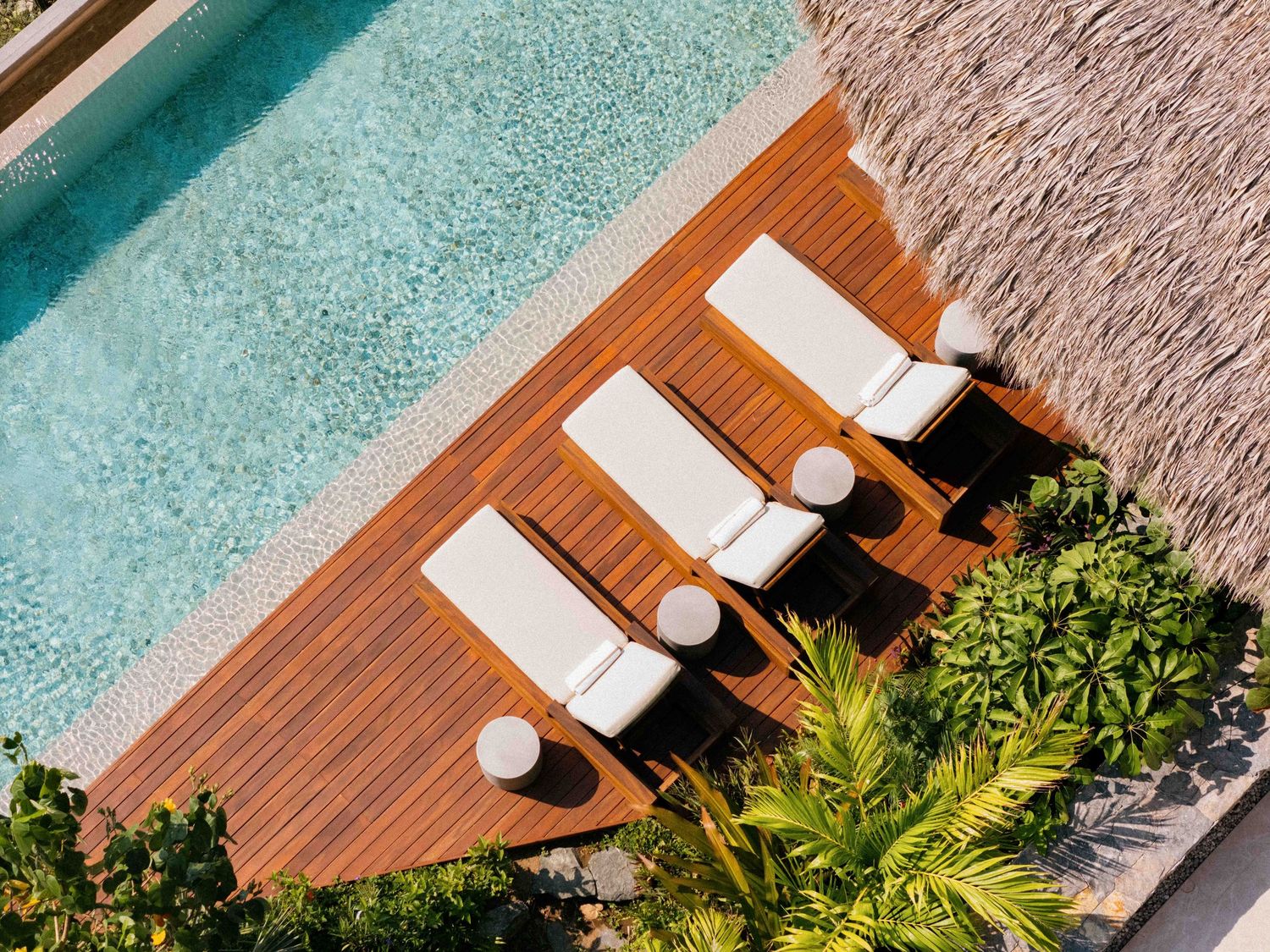 Aerial view of white lounge chairs on wooden deck beside turquoise pool with tropical plants.